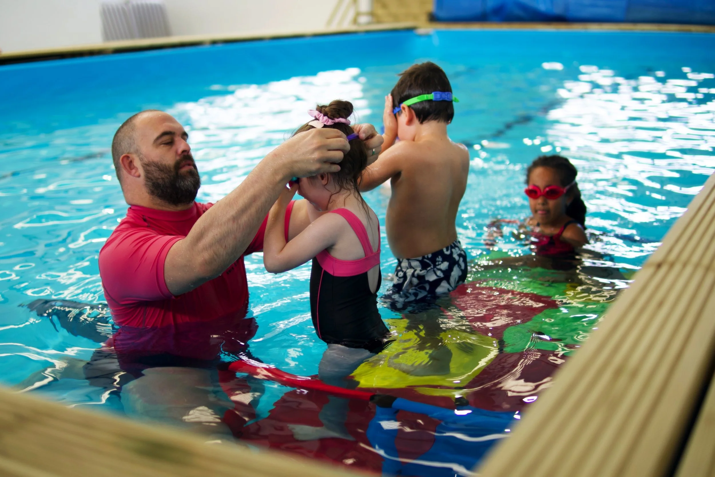 A swimming instructor helping two children put on swim goggles in an indoor pool, with another child swimming in the background.