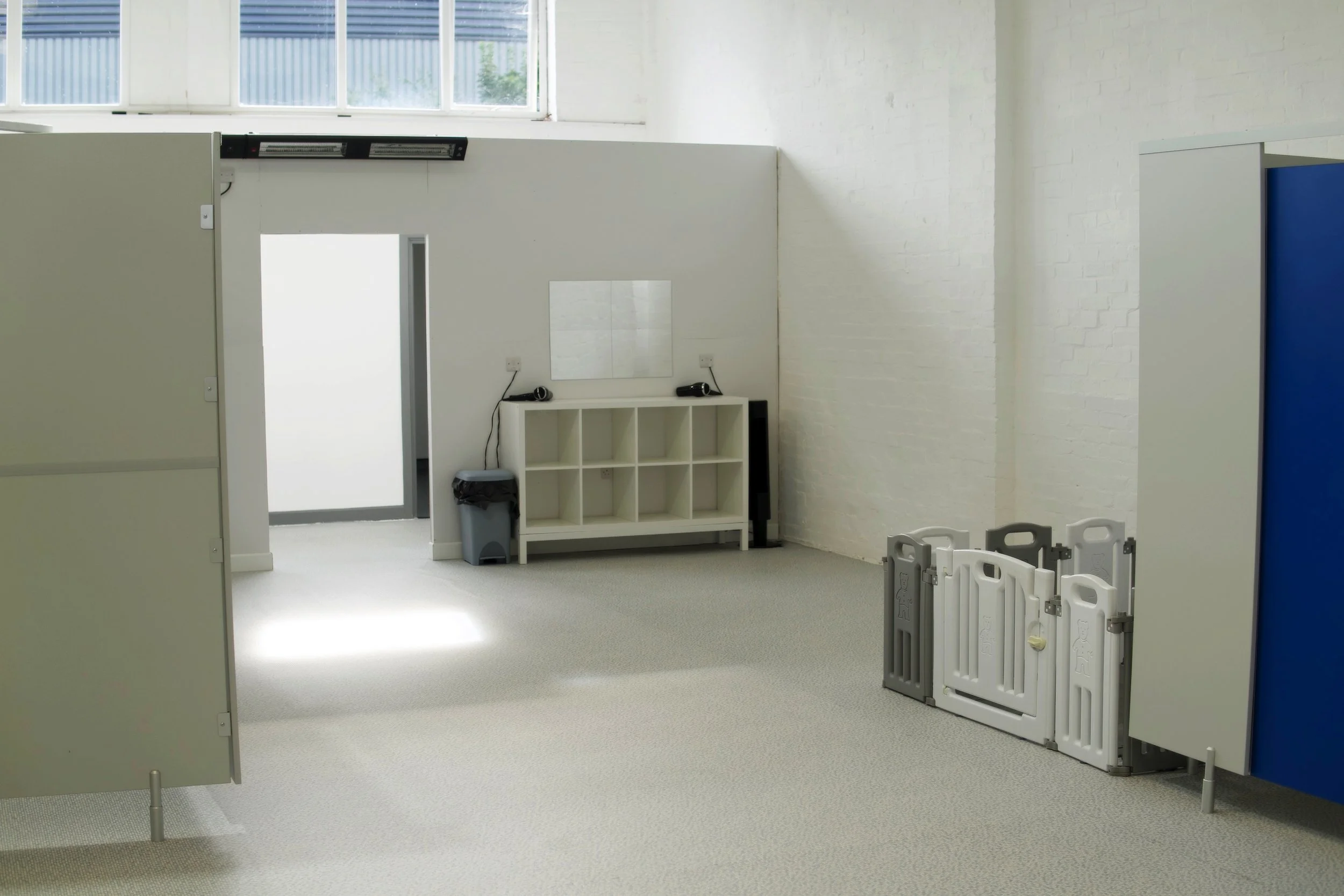 An empty white room with a high window, a white bookshelf with items on top, a gray trash bin, and plastic safety barriers, possibly in a workplace or education setting.