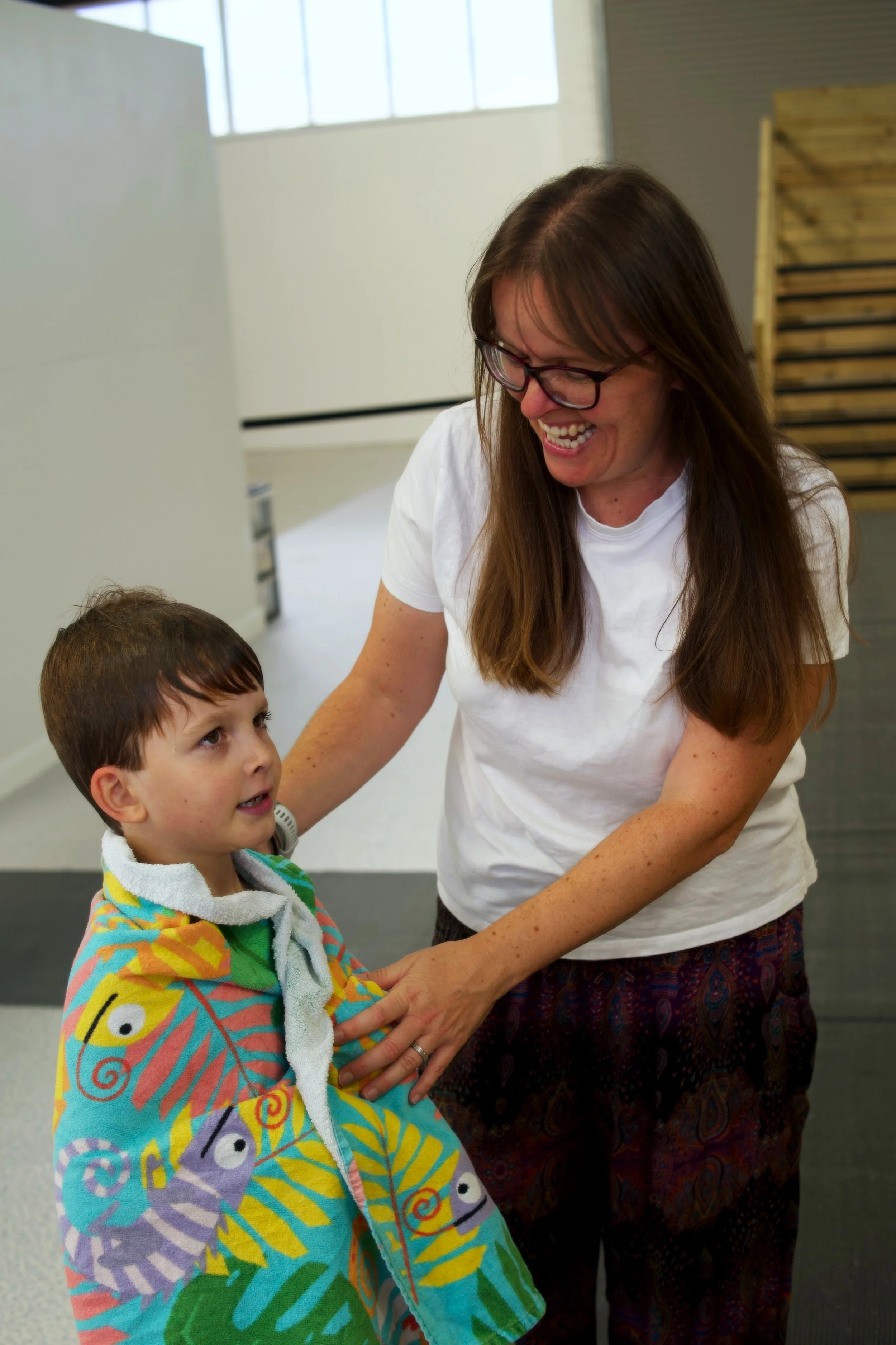 A woman with long brown hair and glasses, smiling, interacts with a young boy. The boy is wrapped in a colorful towel with cartoon characters. They are indoors in a room with large windows and some wooden furniture in the background.