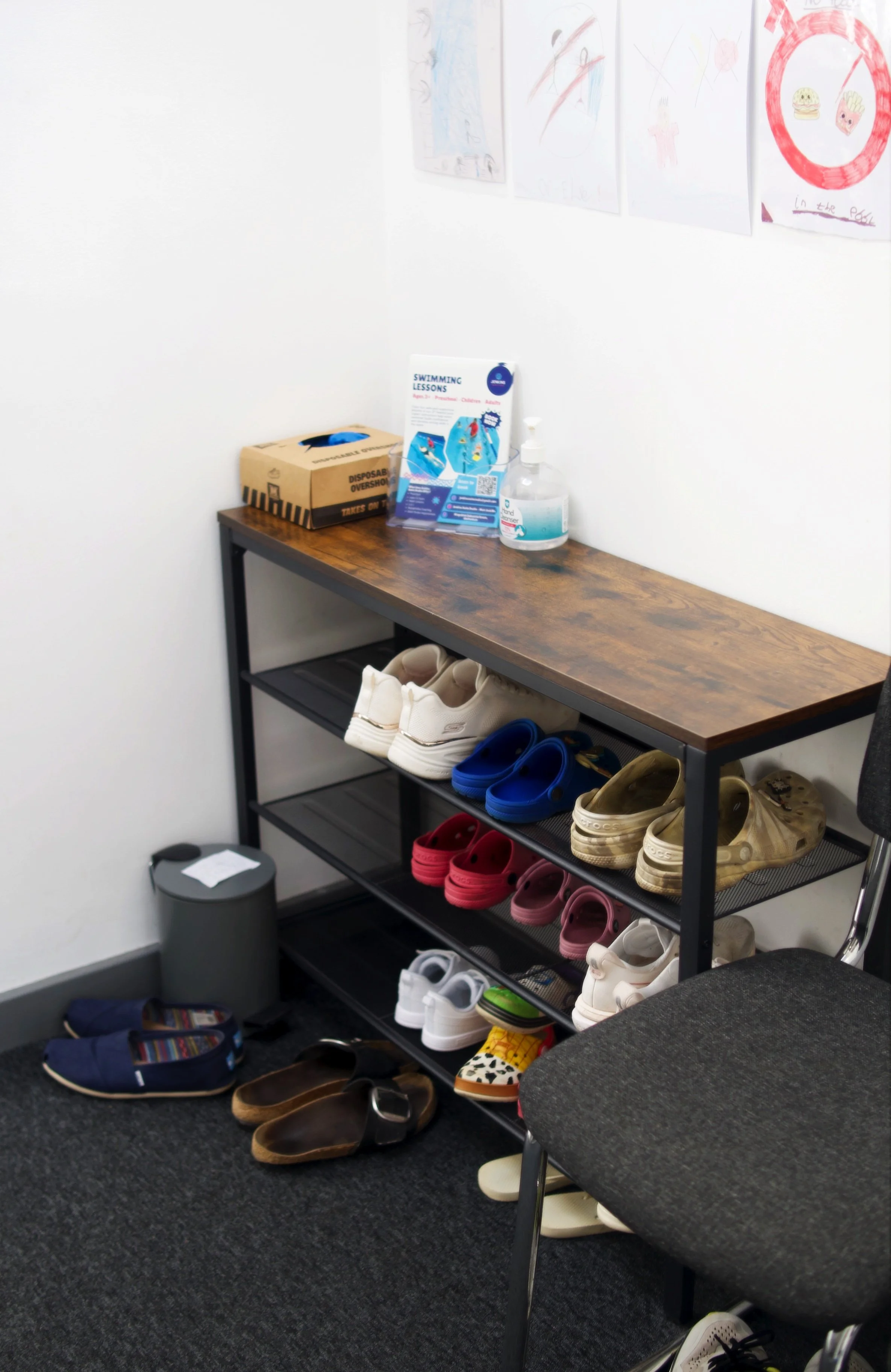 A shoe rack with various shoes and sandals, a pair of white shoes, blue sandals, beige clog shoes, and pink shoes, located in a hallway with a gray carpet. On top of the shoe rack are some children's items, including a swimming lessons flyer and a bottle of hand sanitizer. There is a gray trash bin next to the shoe rack and a gray cushioned chair nearby.