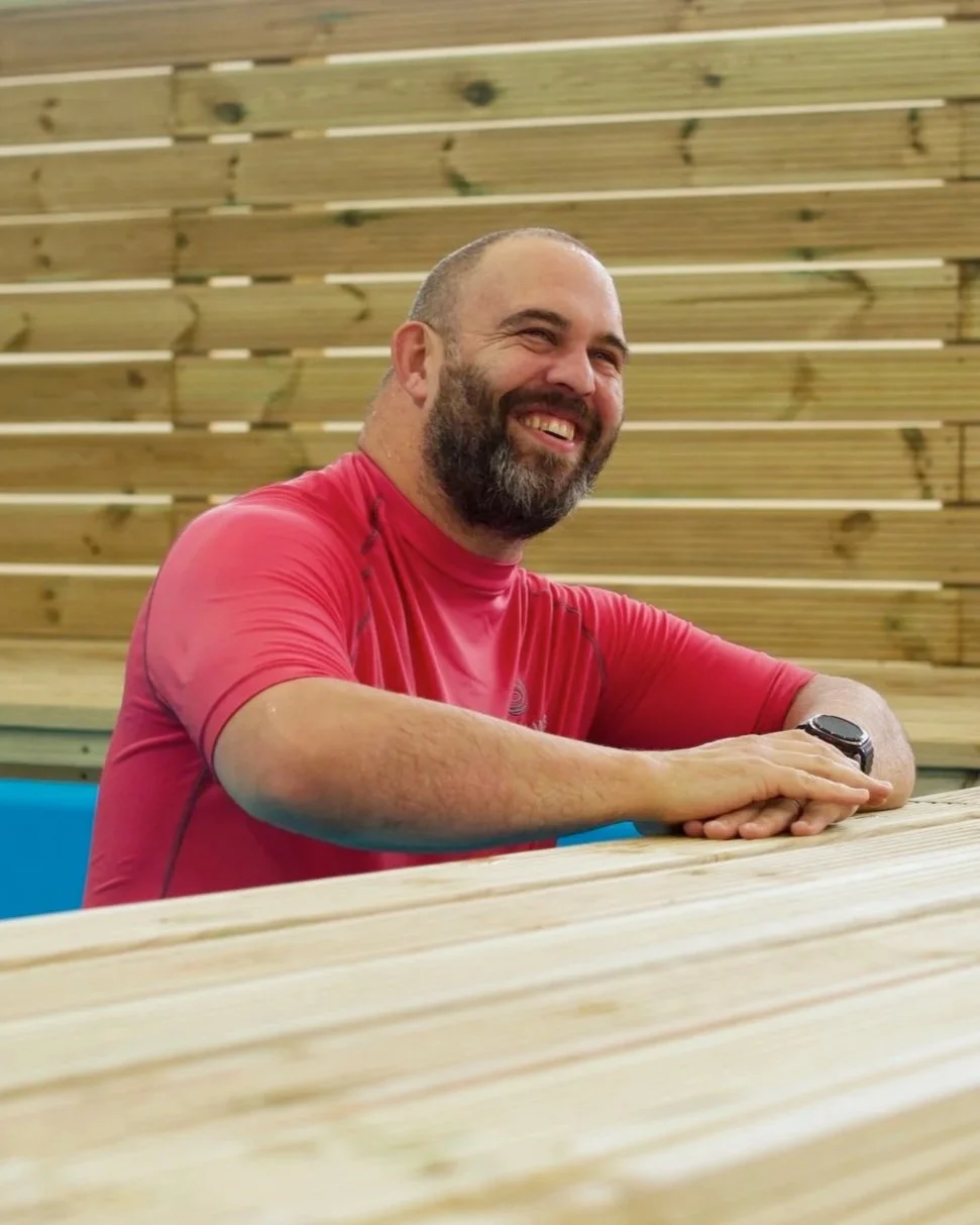 A smiling man sitting at a wooden table in front of a wooden wall, wearing a red athletic shirt and a black smartwatch.