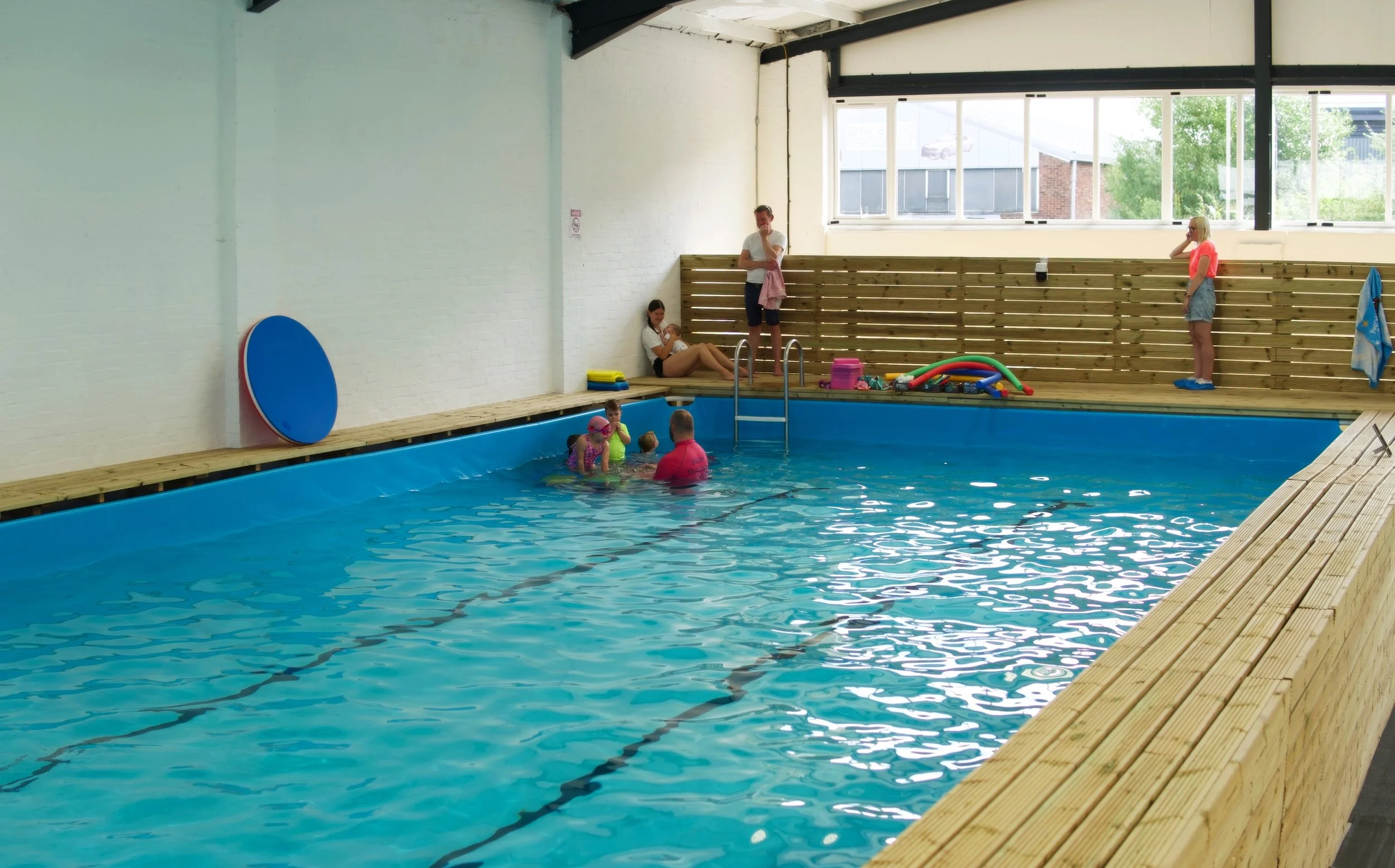 Indoor swimming pool with children enjoying swimming lessons, supervised by a swimming instructor. The pool is surrounded by a wooden deck, with toys and equipment nearby. Bright natural light enters through large windows.
