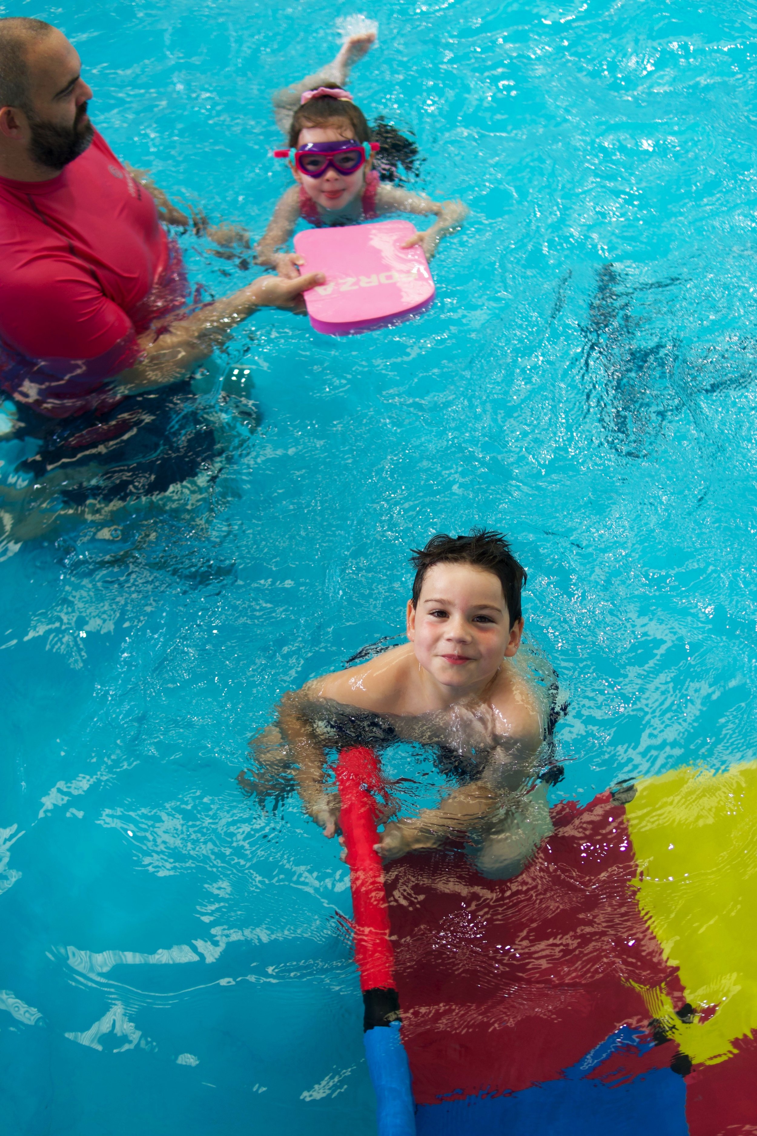 A man helping a young girl with goggles and a swim cap in a swimming pool, holding a pink kickboard. A smiling boy is in the water in the foreground.