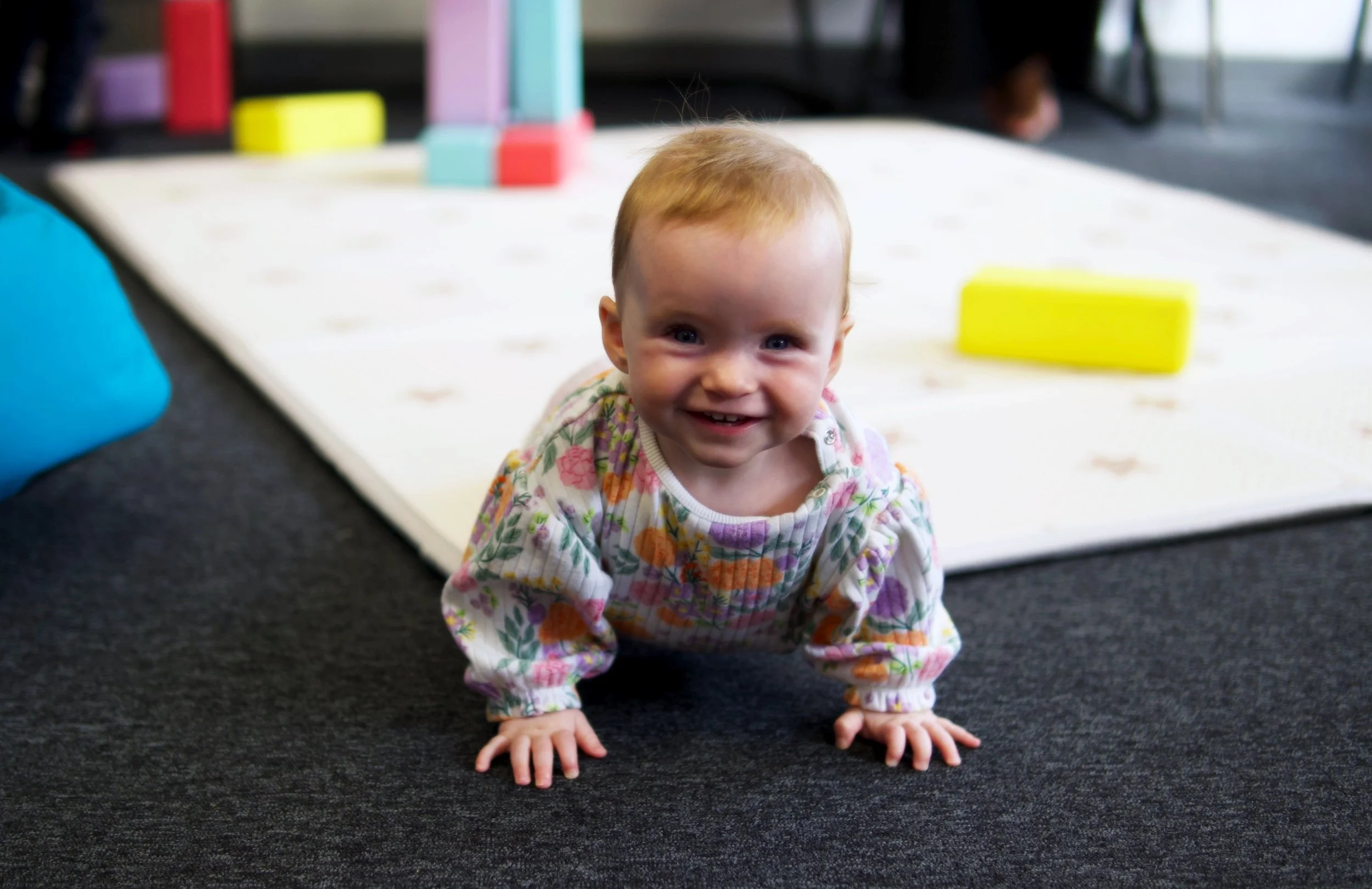A young child with short red hair and blue eyes, smiling and crawling on a black carpeted floor in an indoor setting, wearing a colorful floral-patterned long-sleeve outfit, with a white mat and foam blocks in the background.
