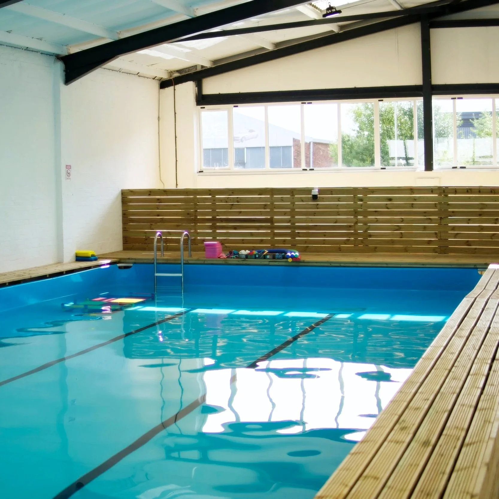 Indoor swimming pool with a wooden deck, blue water, and large windows letting in natural light.