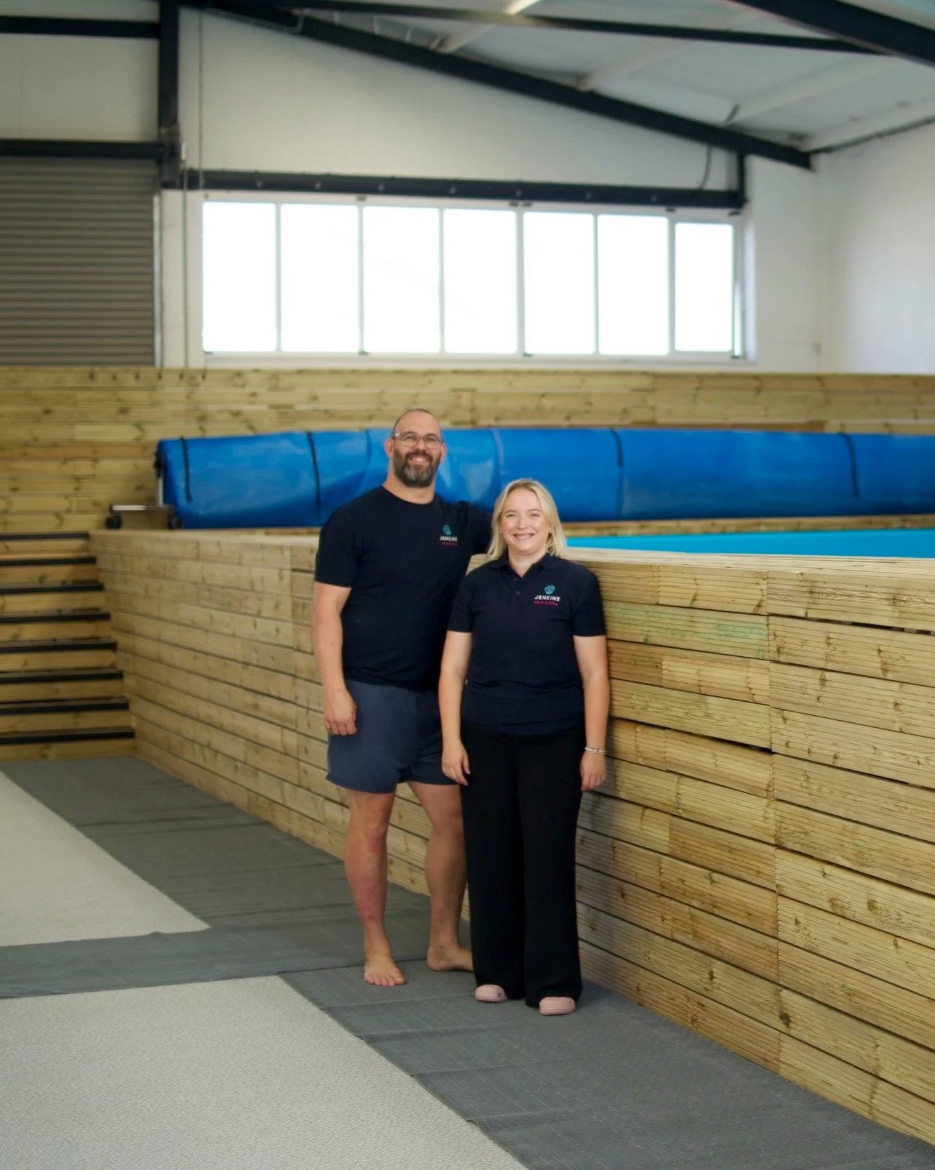 A man and woman standing inside a gym or fitness facility with a wooden stack wall behind them and large windows above. The man is barefoot, wearing a black T-shirt and shorts, and the woman is wearing a black uniform with pink slippers.