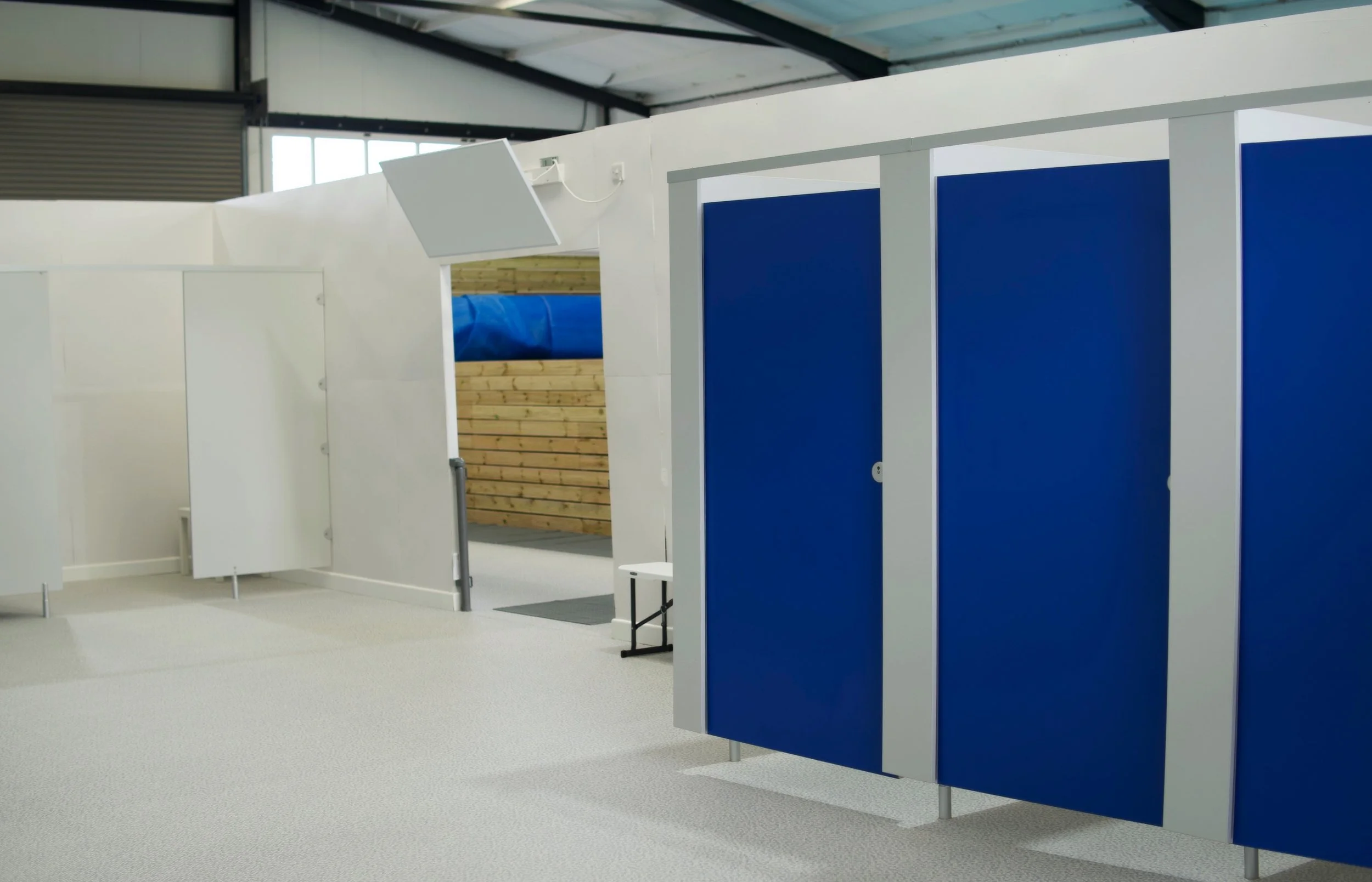 Row of blue and white changing stalls inside a gym or swimming pool facility with a wooden climbing wall in the background.