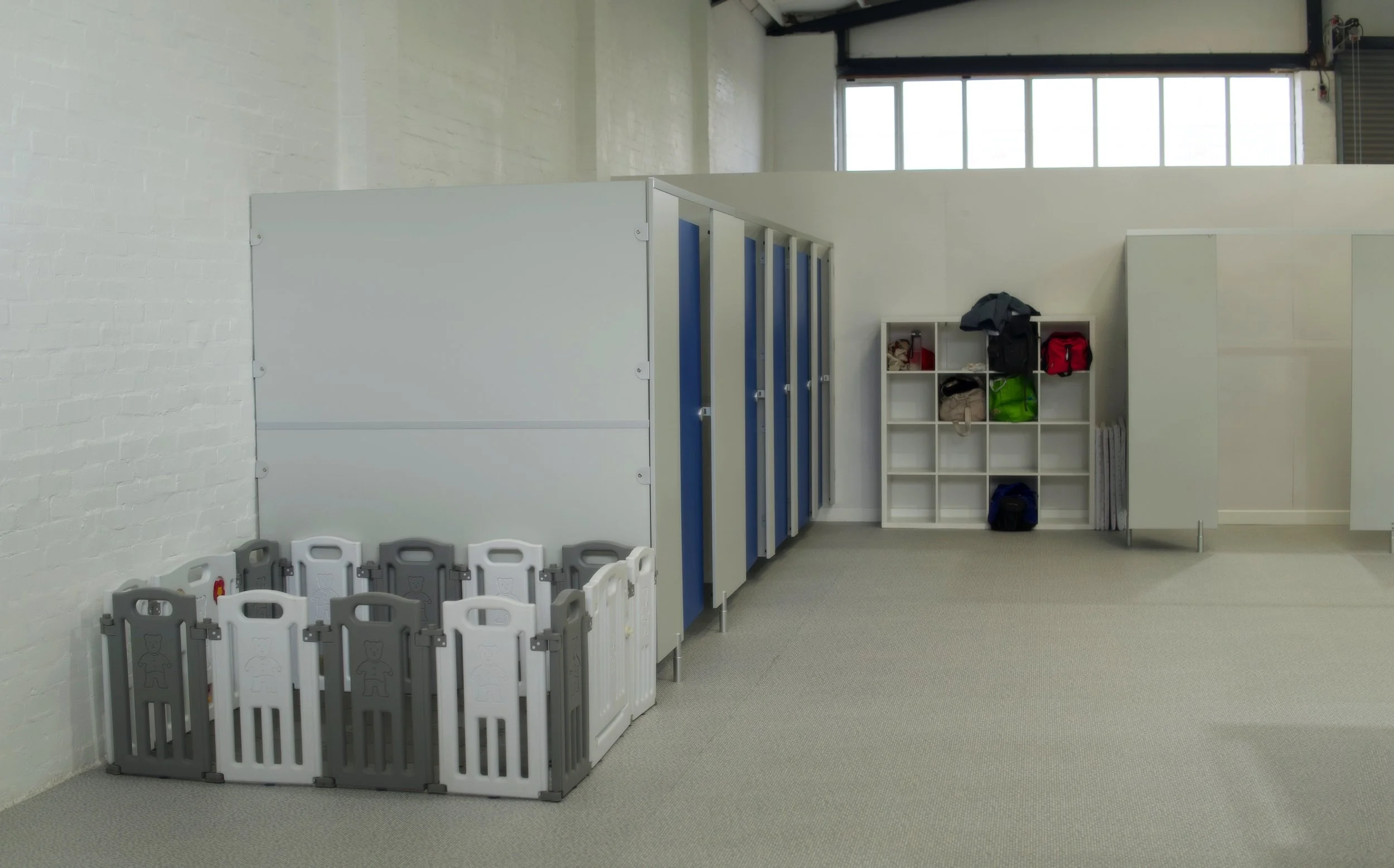 Indoor view of a white brick wall with a row of gray and white baby safety gates in the foreground, lockers on the right with bags on top, and a cubby storage unit with bags and items, all in a well-lit room with large windows.