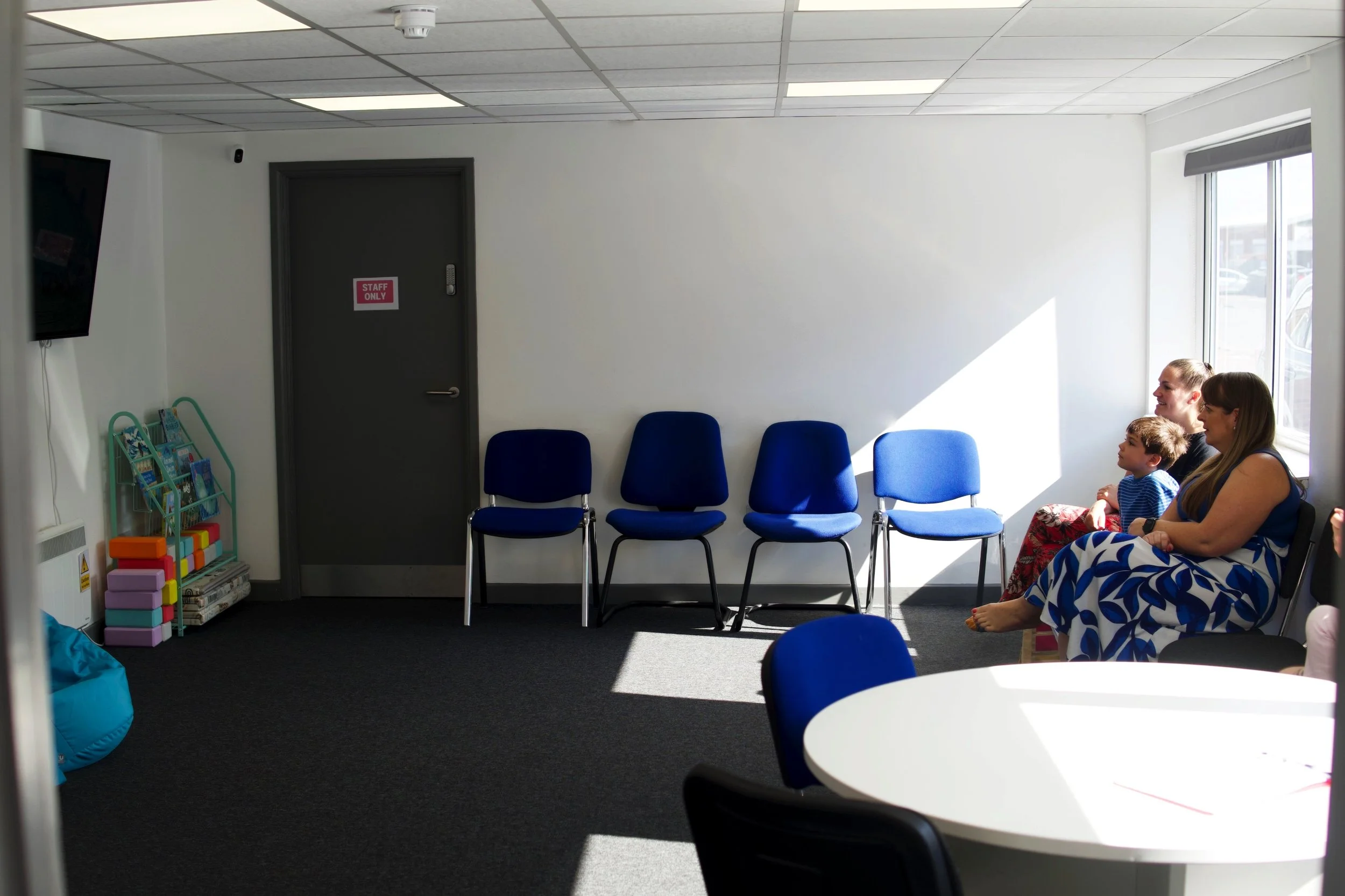 Waiting room with five blue chairs, a window with sunlight, a 'Staff Only' door, a TV, and three people sitting on chairs near the window.