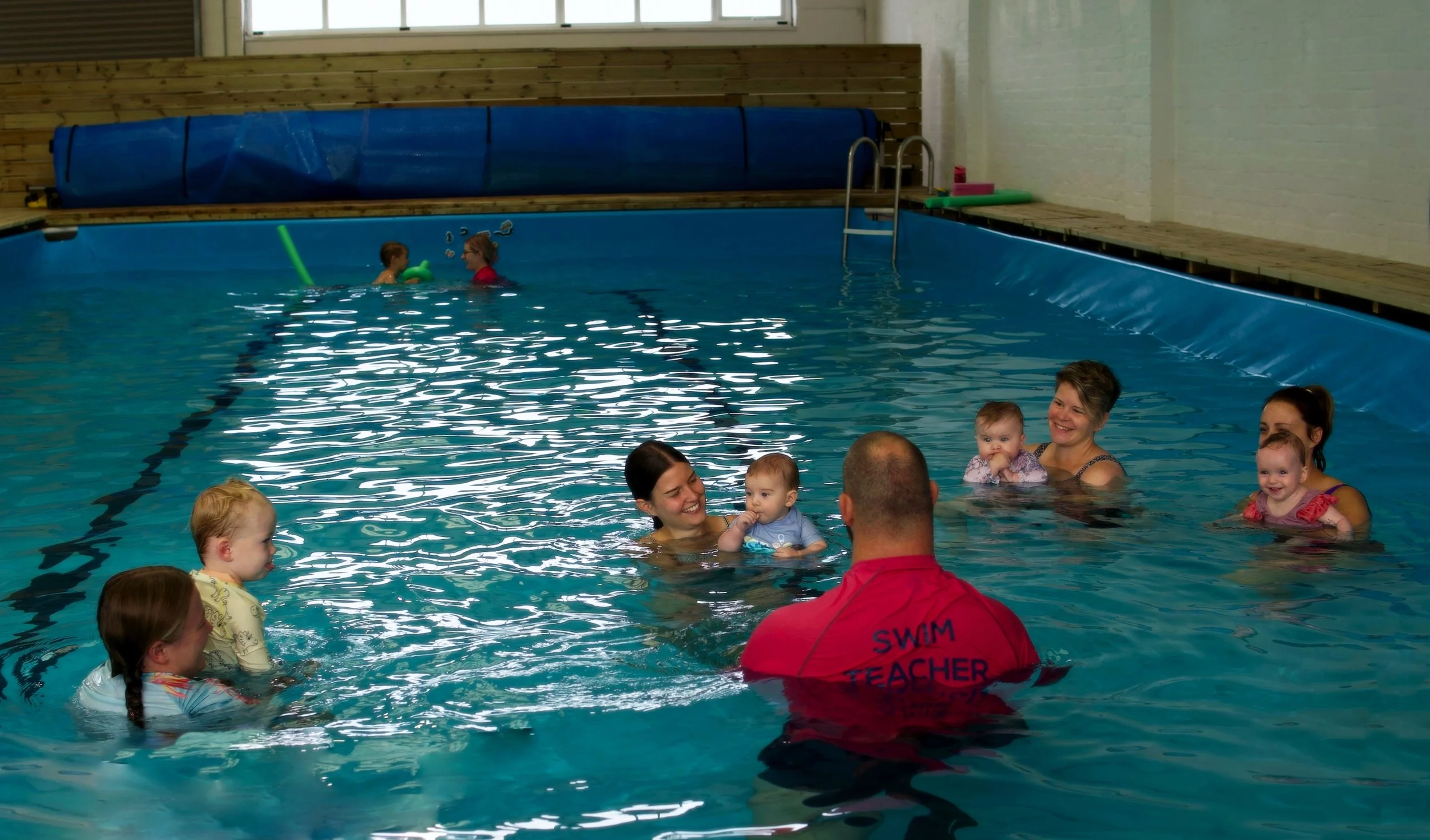 Children and adults in a swimming pool participating in a swimming lesson, with an instructor wearing a red shirt that says 'SWIM TEACHER' on the back.