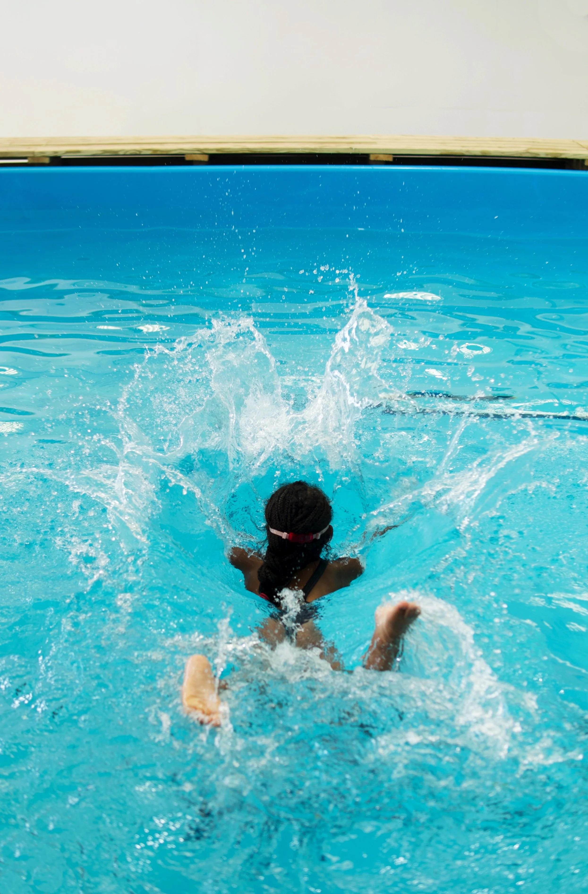 A young girl swimming underwater in a pool, with her head submerged, arms extended forward, and legs kicking behind. Water splashes around her as she swims.