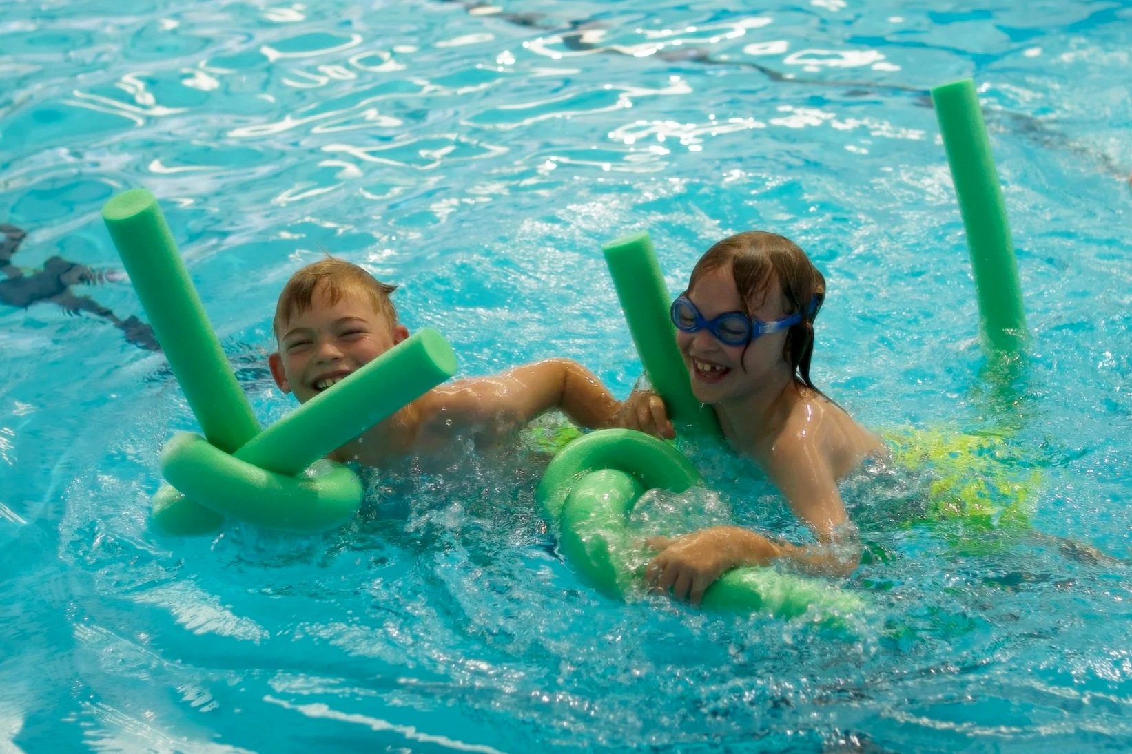 Two children happily playing with pool noodles in a swimming pool, smiling and enjoying the water.