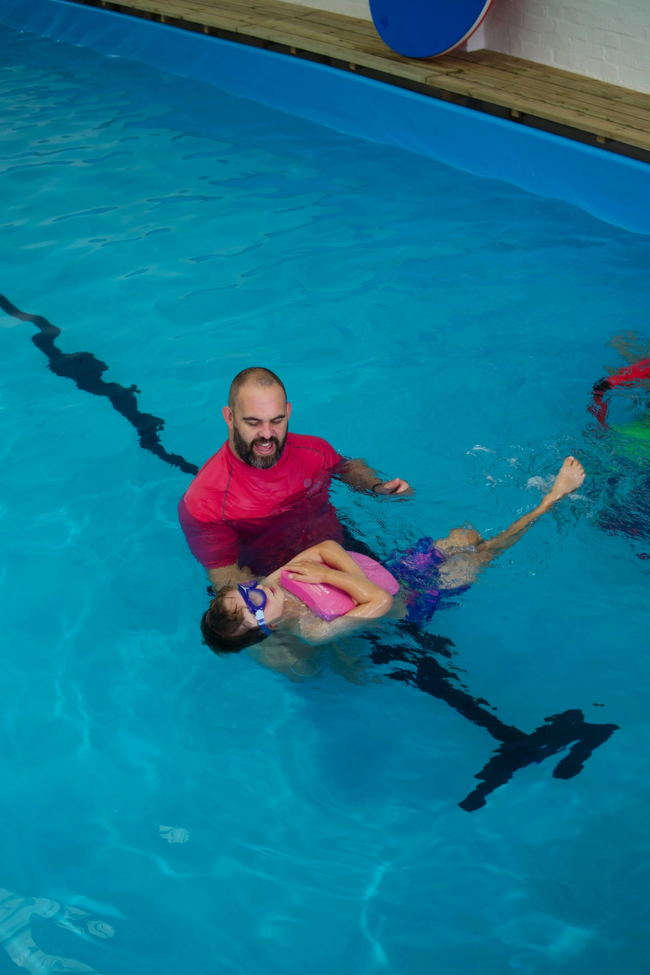 A man and a young girl are in a swimming pool. The man, wearing a red shirt, is supporting the girl, who is wearing blue goggles and holding a pink floaty, as she swims in the pool.