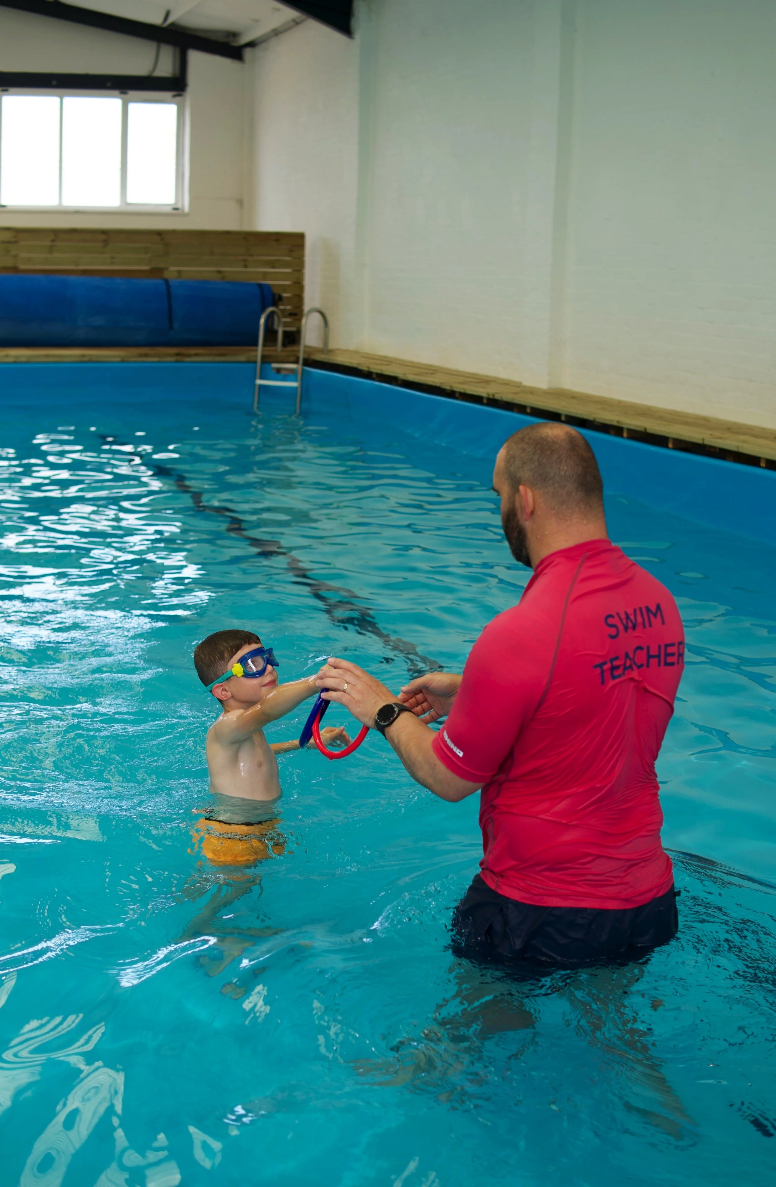 A boy with swimming goggles is being handed a lifebuoy ring by a swim instructor in an indoor swimming pool. The instructor is wearing a red shirt that says 'Swim Teacher'.