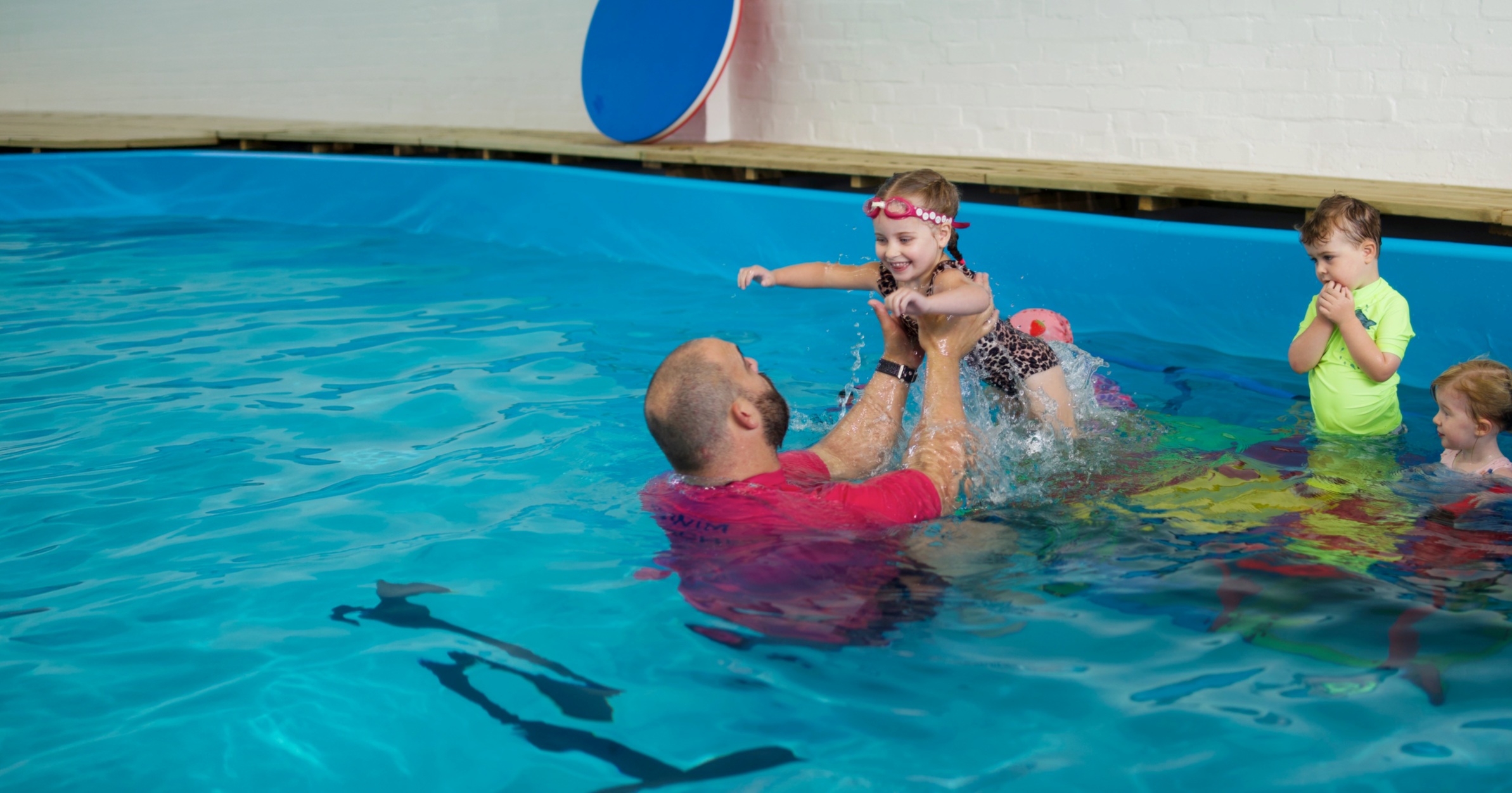Children and an instructor in a swimming pool, with the instructor lifting a young girl wearing pink goggles and a leopard print swimsuit, while three other children watch.