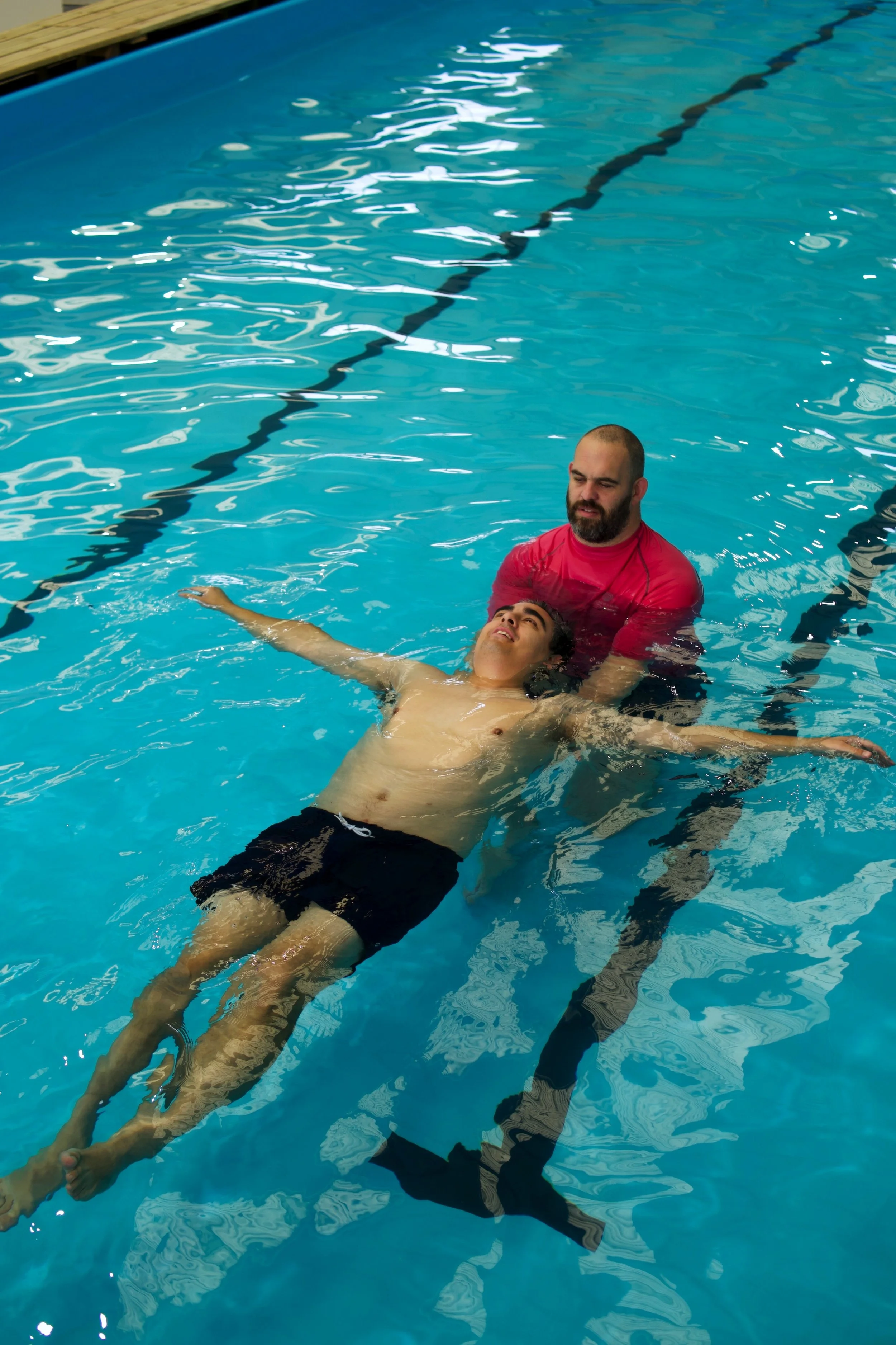 A man is holding and supporting another man in a prone position in a swimming pool. The man being held appears to be unconscious or in distress, with his eyes closed and arms extended outward. The swimmer is wearing a red shirt, and the person being supported is shirtless with black shorts.