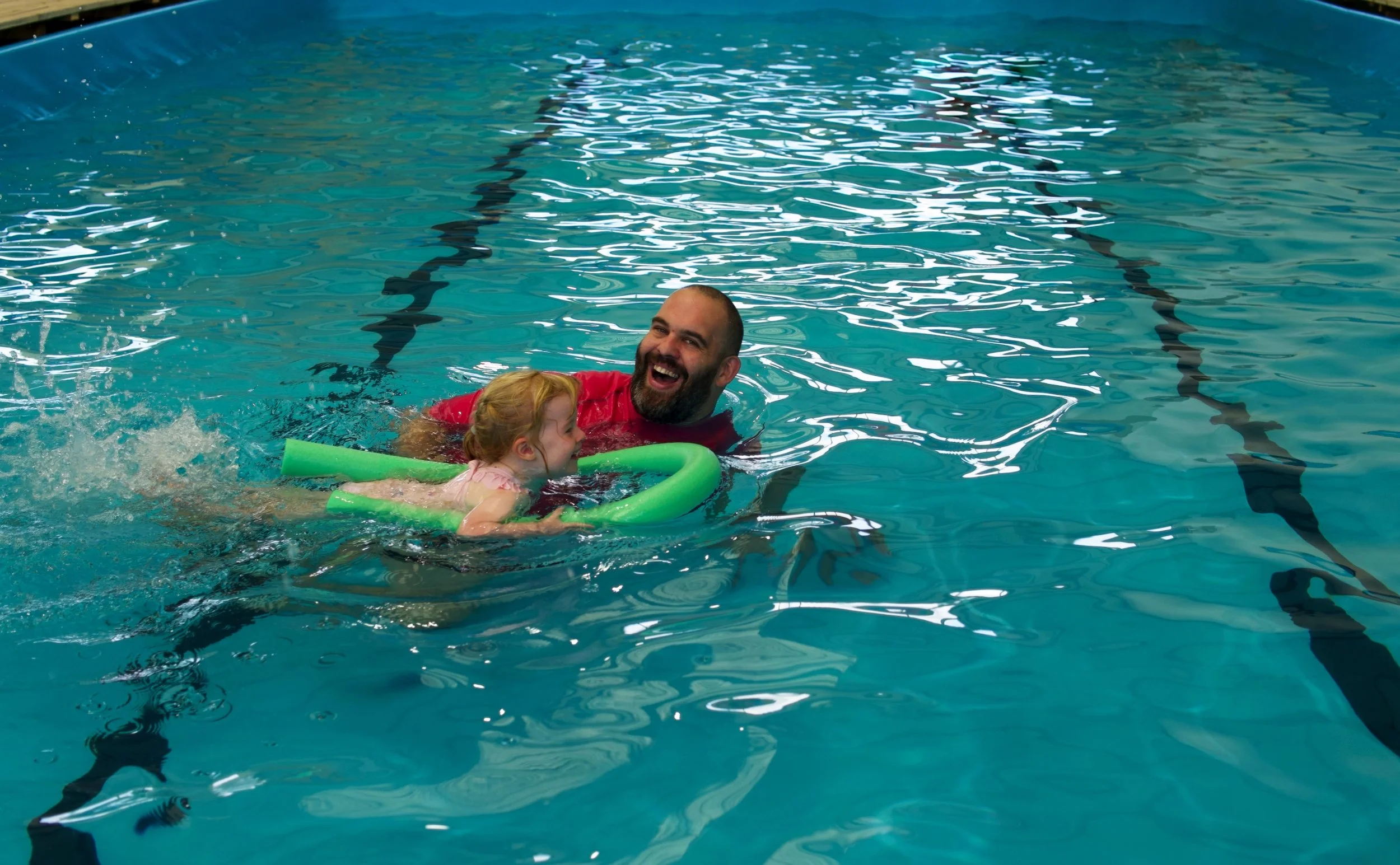 A man and a young girl swimming in a pool, with the man holding the girl using a green pool noodle, both smiling and enjoying themselves.