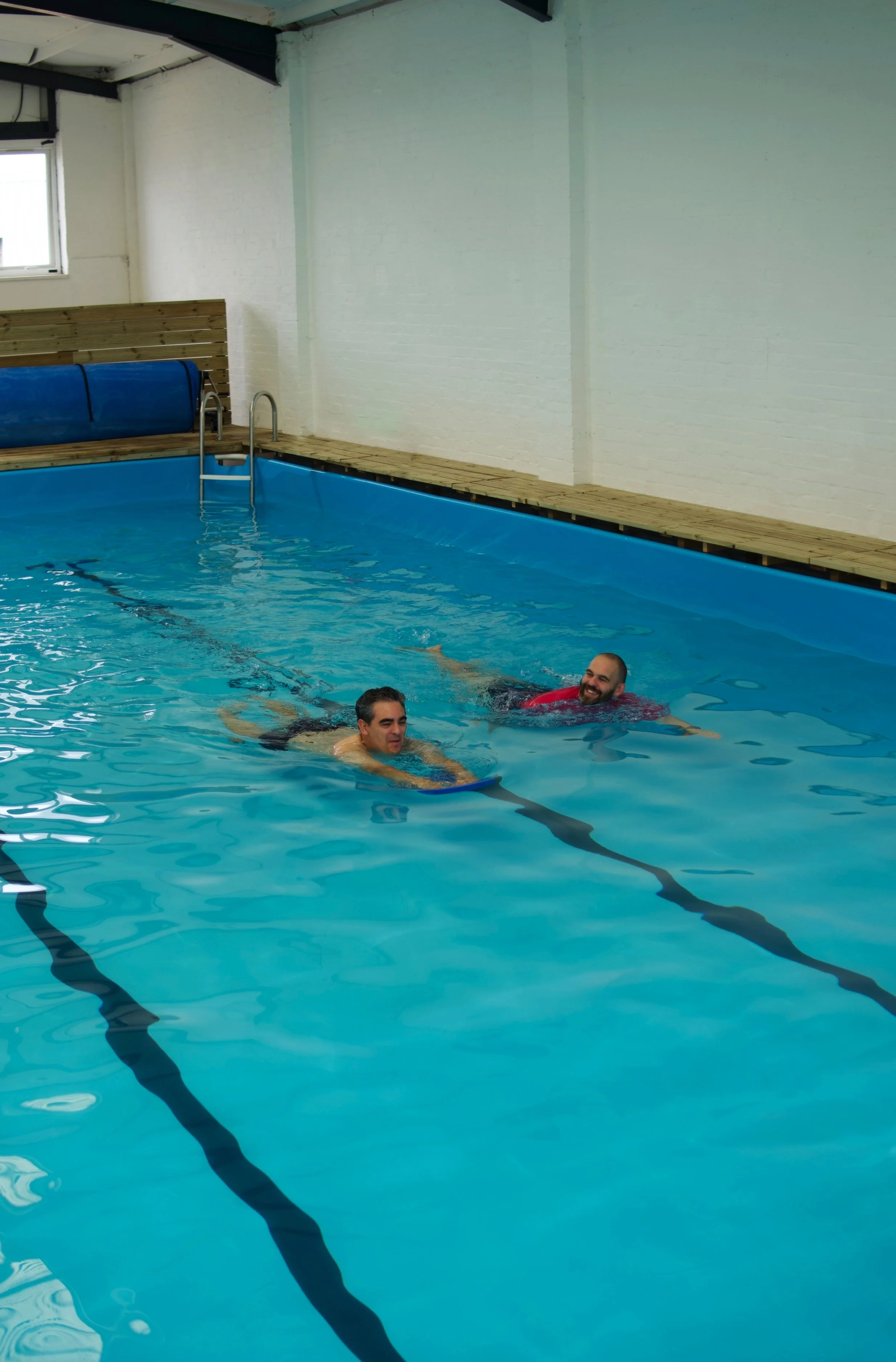 Two men swimming in an indoor swimming pool with a wooden deck on the side and a ladder, white brick walls, and a small window.