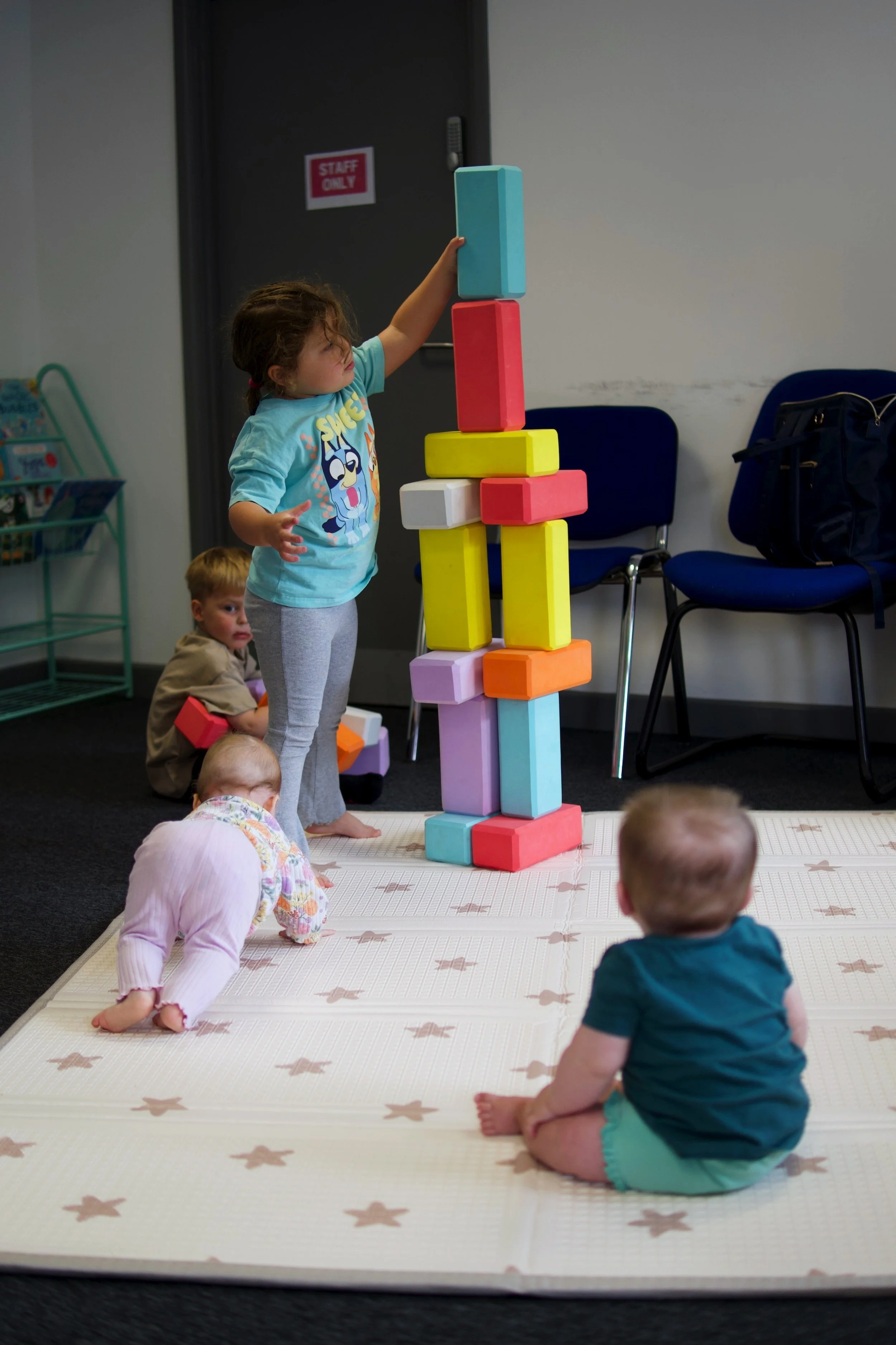 A young girl stacking large foam blocks on top of each other during a play session, with several other children sitting or crawling on a play mat in the foreground, in a classroom or playroom setting.