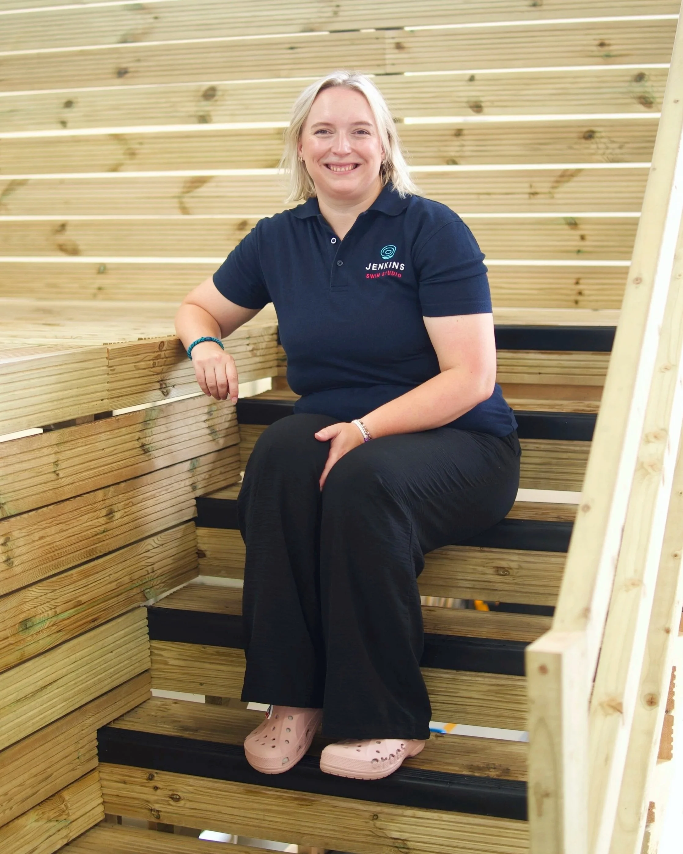 A woman with blonde hair smiling, sitting on wooden stairs inside a building, wearing a navy polo shirt with a logo and the text "Jenkins Swim Studio," black pants, and pink Crocs.
