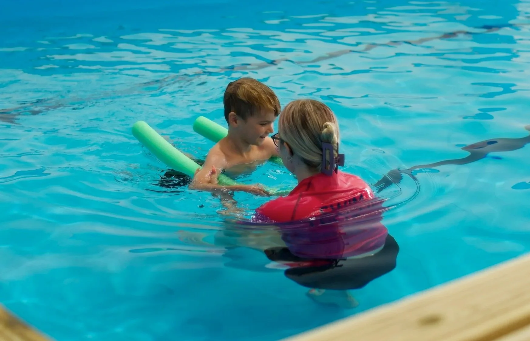A young boy with brown hair in swim trunks learning to swim with a female instructor in a swimming pool. The instructor, wearing glasses and a red life jacket, is supporting the boy who has green pool noodles under his arms.