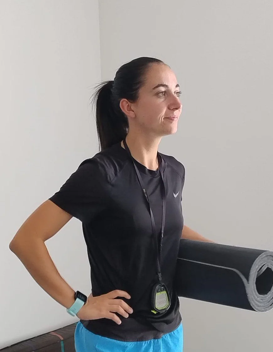Une femme coach sportive en tenue de sport noire et bleue avec une montre et un chronomètre autour du cou, tenant un tapis de fitness.
