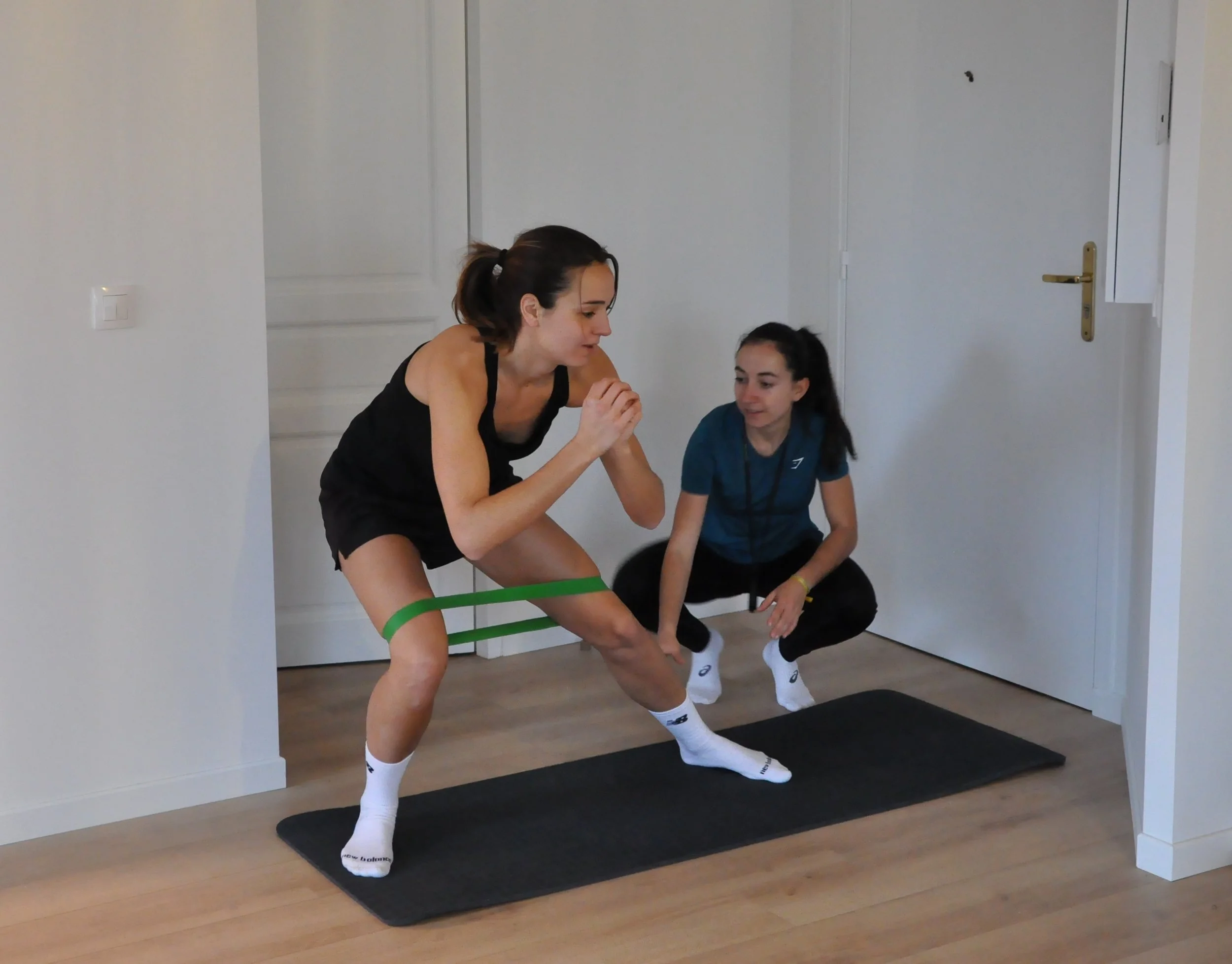 Une femme et une coach s'entraînent avec une bande élastique dans une pièce, portant des chaussettes de sport et des vêtements d'entraînement.