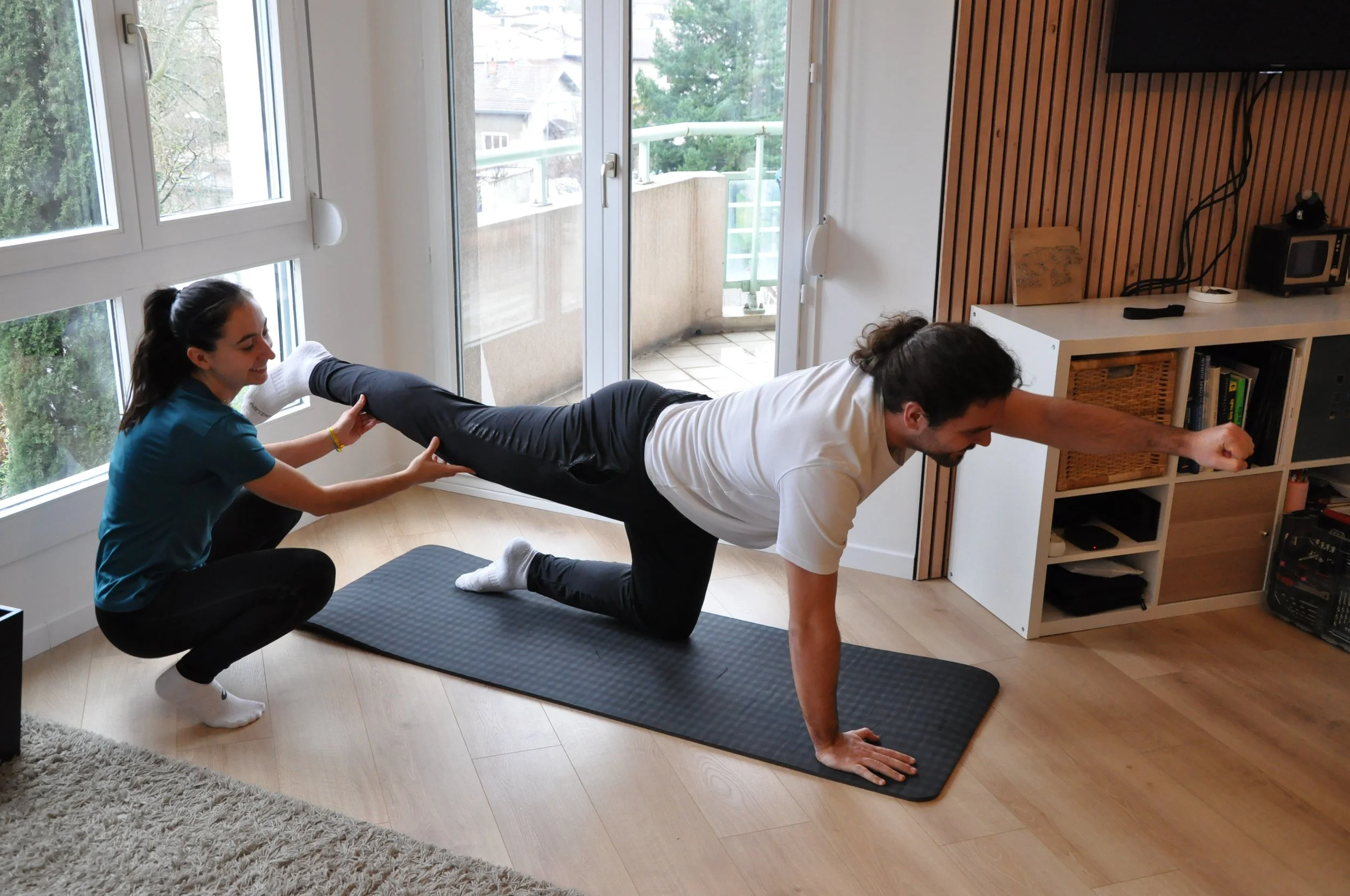 Deux personnes pratiquant du sport dans un intérieur, avec un homme en position de planche et une femme coach aidant à soutenir ses jambes, sur un tapis de sport noir.