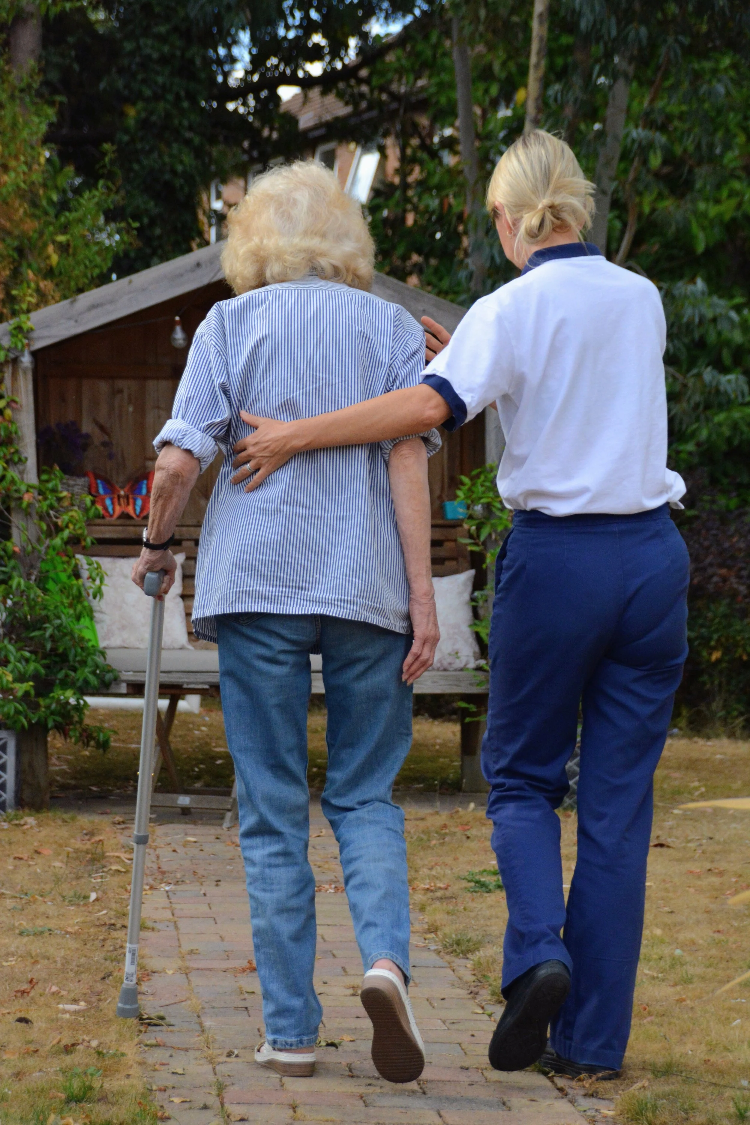 An elderly woman with white hair using a cane walk arm in arm with a young caregiver with blonde hair in a low bun, in a backyard garden with trees and greenery.