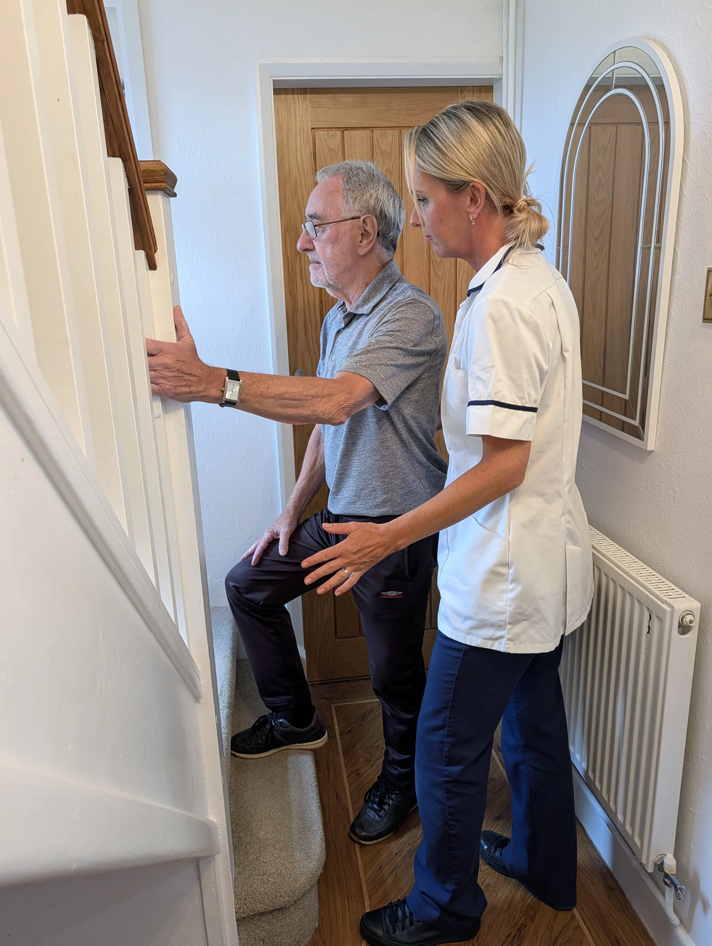 A man sitting on a step with his leg elevated, receiving care or examination from a woman in a white medical uniform, in a room with wooden doors, a radiator, and a mirror on the wall.