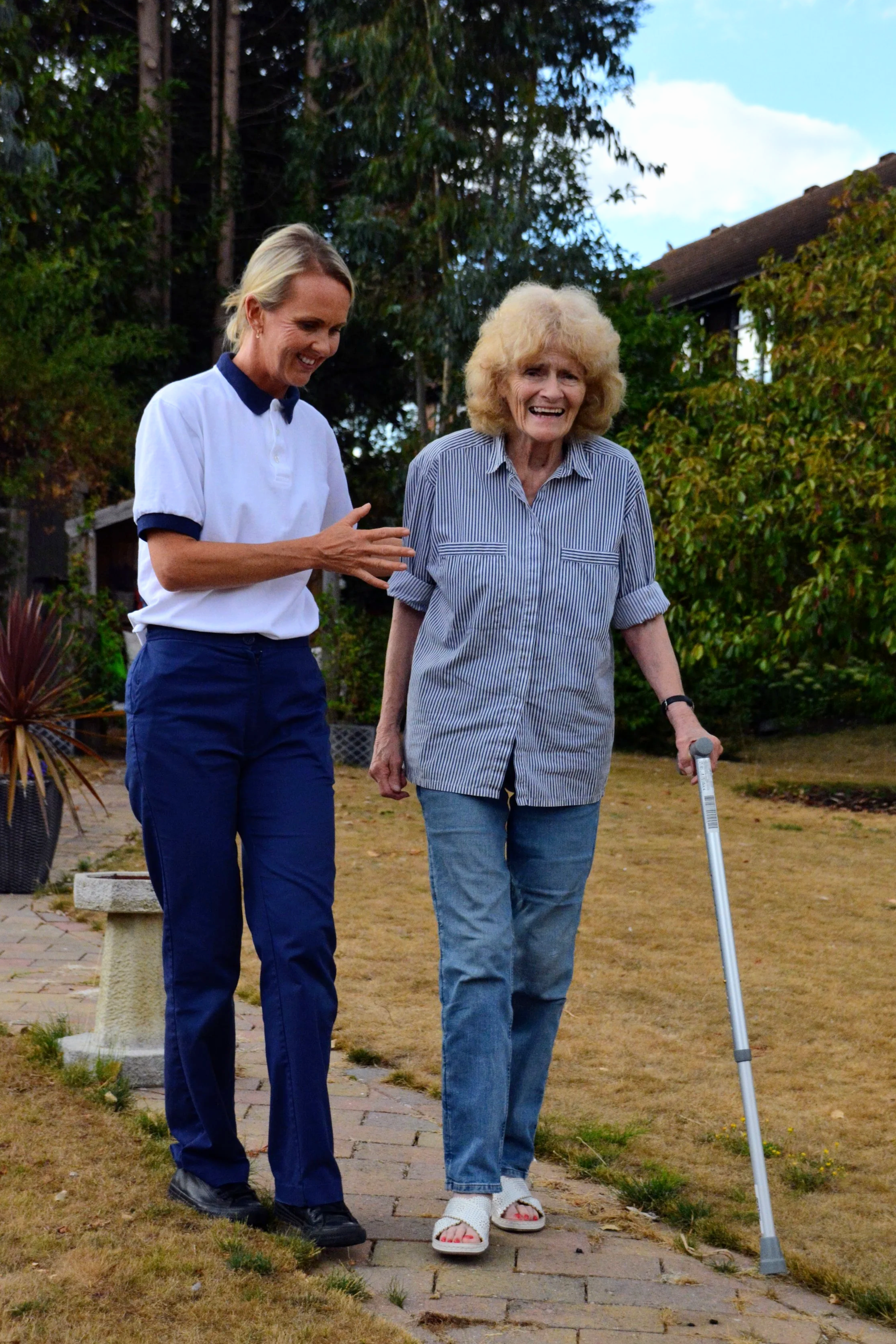 Two women walking outdoors on a brick pathway. One woman is elderly with curly blonde hair, using a walking cane, and wearing a striped shirt and jeans. The other woman is younger with short blonde hair, wearing a white polo shirt with navy edges and navy pants. They are smiling and talking, surrounded by greenery and a house in the background.