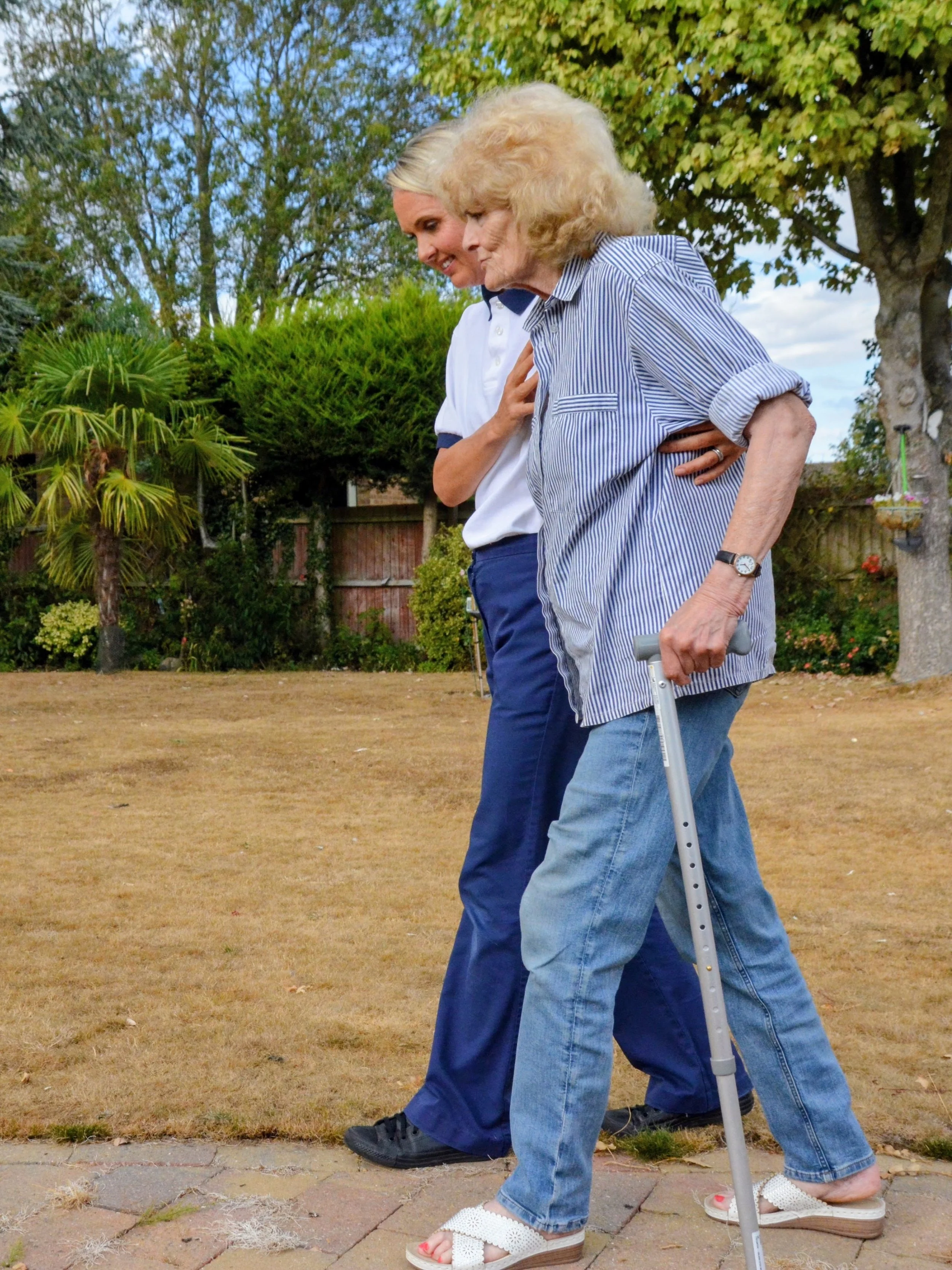 Two women walking outside, one elderly with a cane and the other younger, closely walking together on a sunny day in a garden or park area with trees and bushes.