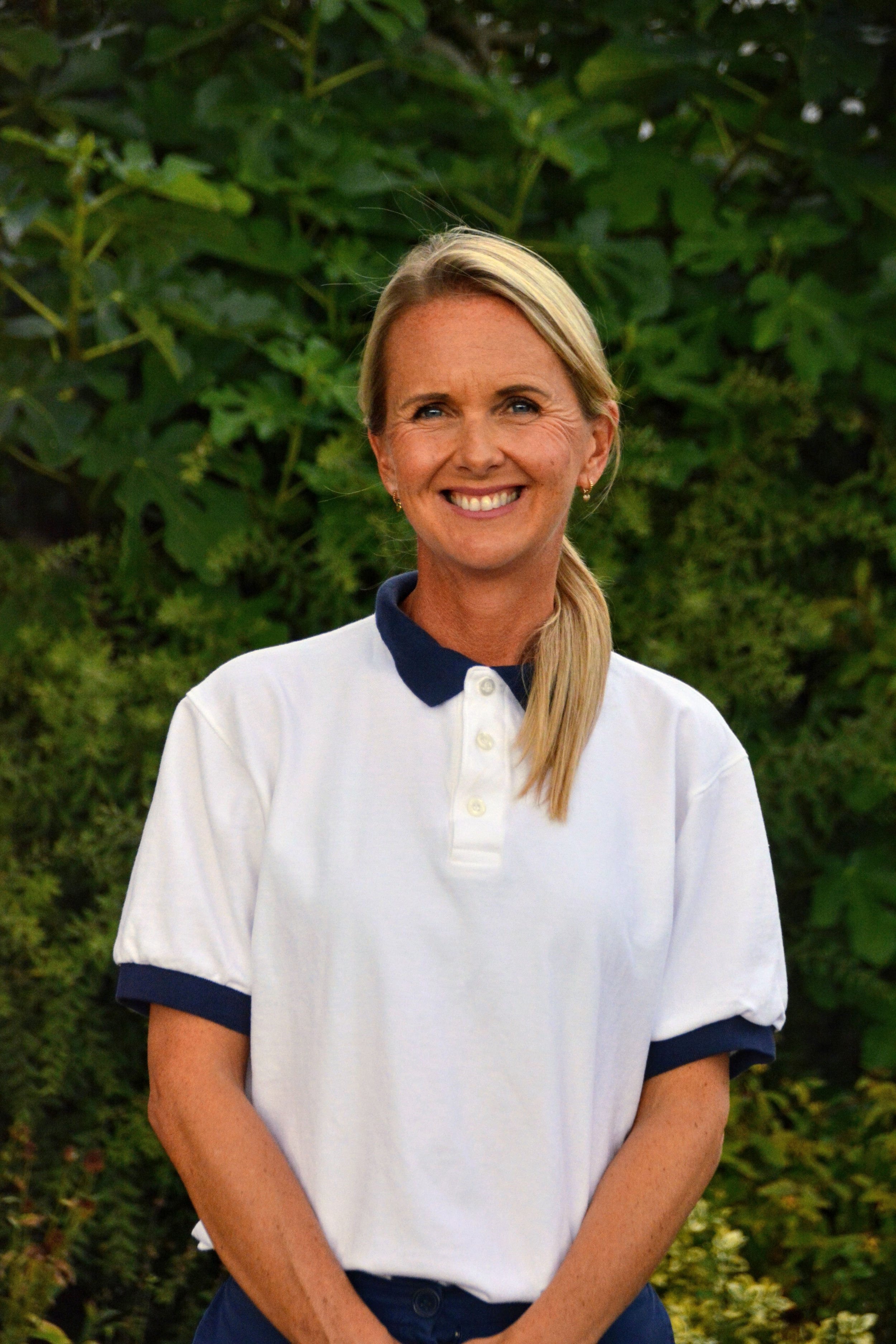 A woman with blonde hair tied back, wearing a white polo shirt with navy collar and sleeve trim, smiling outdoors with greenery in the background.