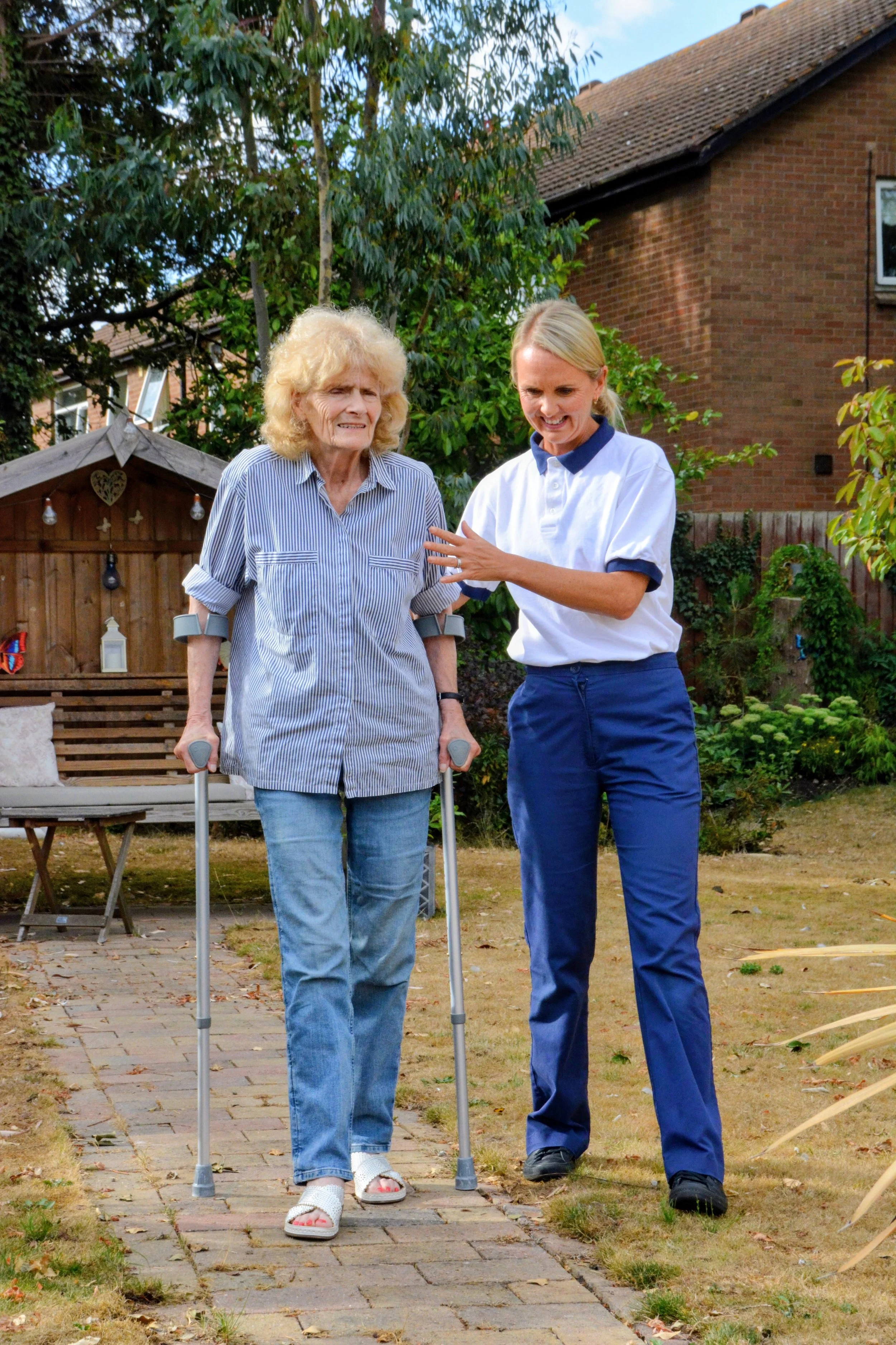 A young woman helping an elderly woman walk with crutches in a backyard garden.