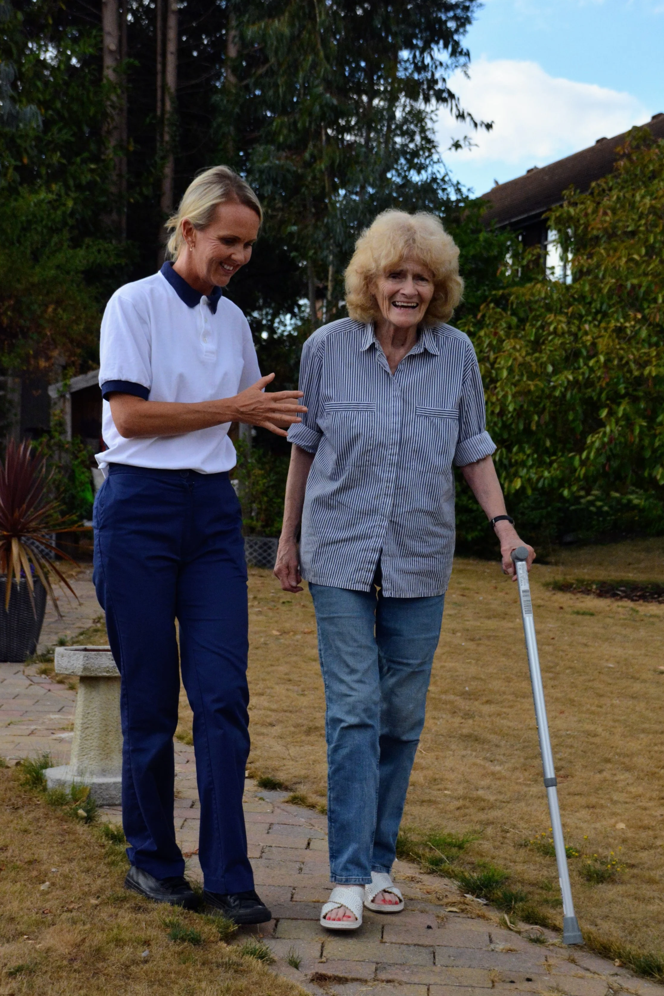 A woman walking in a garden with a cane, accompanied by a caregiver or friend, during the daytime. The woman is smiling, wearing a striped shirt and sandals, and the caregiver is smiling and wearing a white polo shirt and dark blue pants.