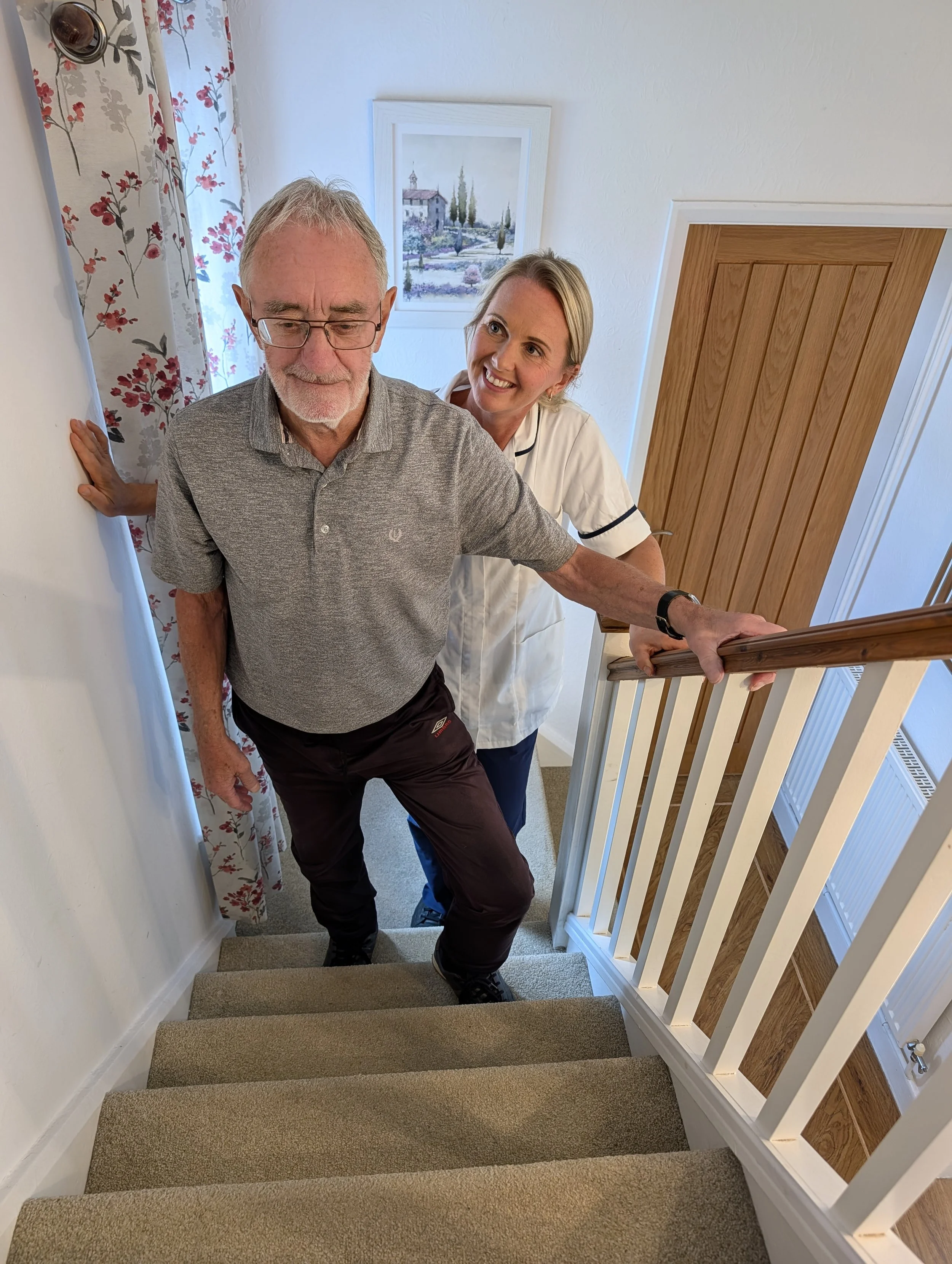 An elderly man with glasses and a gray polo shirt is descending a carpeted staircase, supported by a cheerful woman with blonde hair, wearing a white blouse. The setting appears to be a cozy home interior with artwork on the wall and floral curtains.