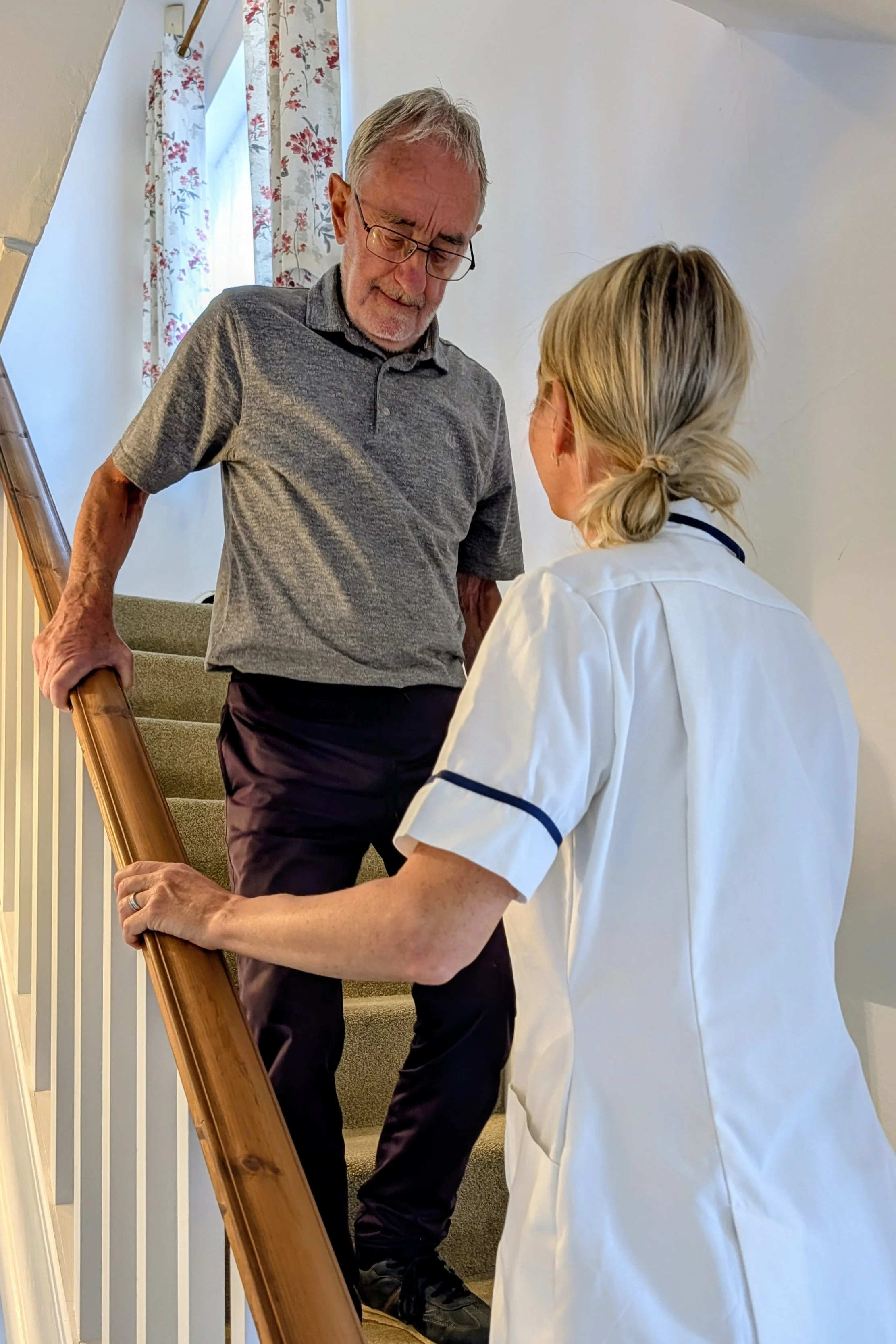 An elderly man with gray hair and glasses standing on a staircase, holding the handrail, while a woman in a white uniform with dark trim kneels in front of him, engaging in conversation. The setting is a home interior with floral curtains and a plain wall.
