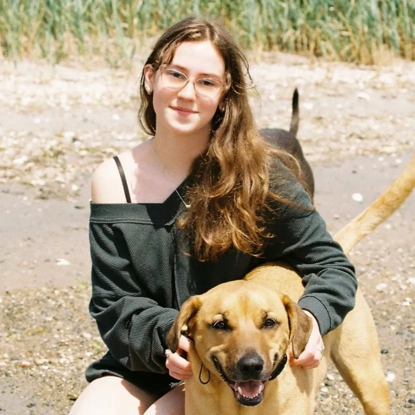 A young woman with long brown hair, wearing glasses and a black off-shoulder top, sitting outdoors with a happy tan dog.