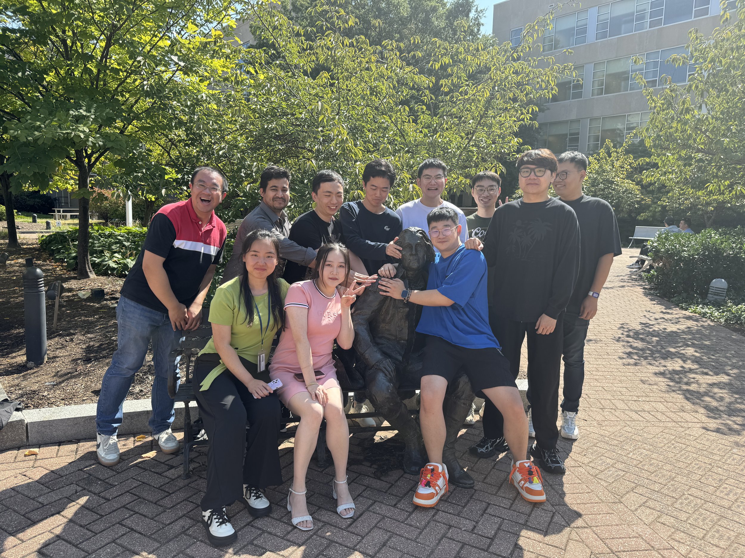 Group of diverse young adults posing outdoors around a statue of a George Washington, with trees and a modern building in the background.