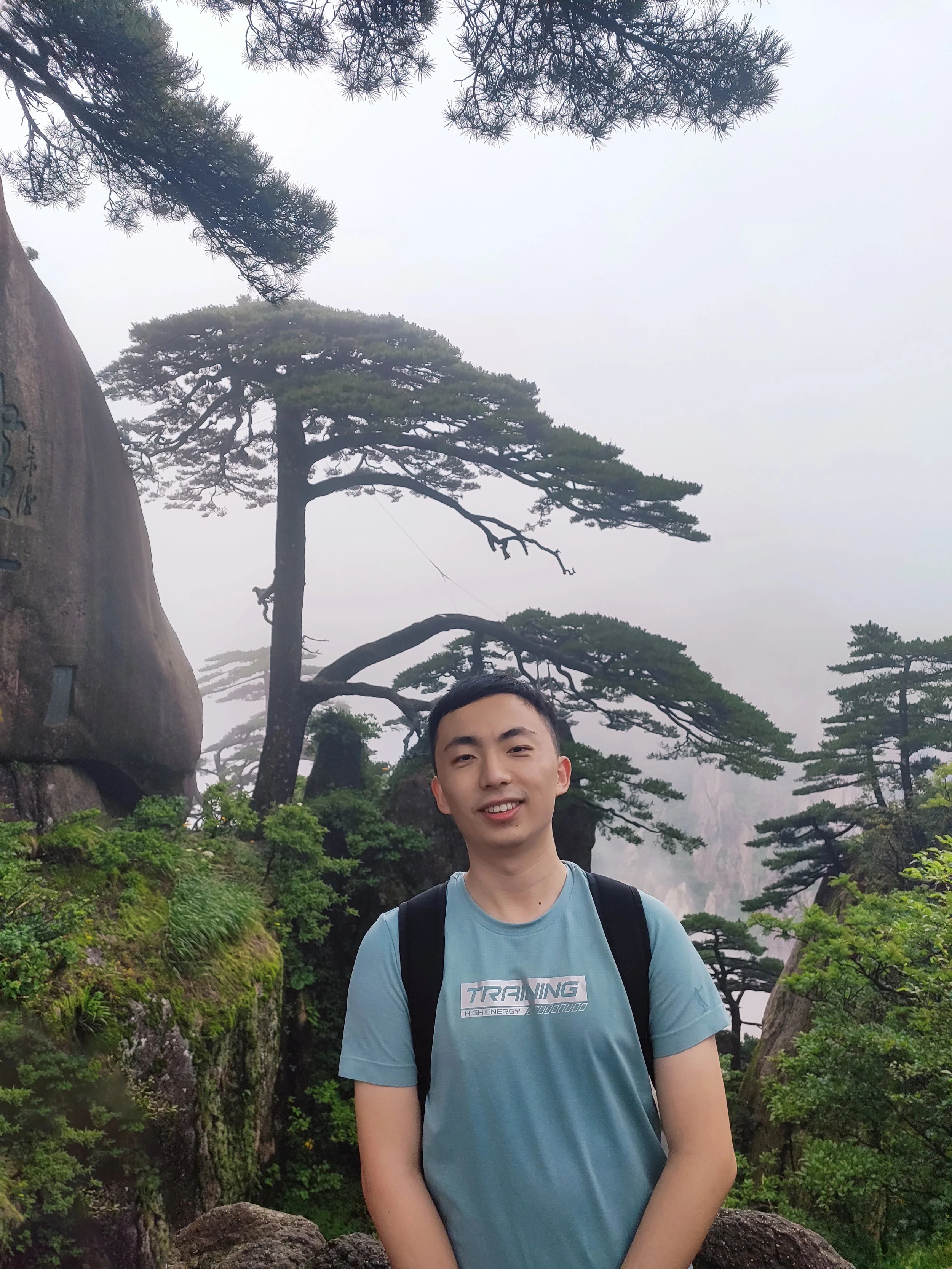 Young man with short dark hair smiling in front of a lush, mountainous landscape with tall, twisted pine trees and misty mountains in the background.