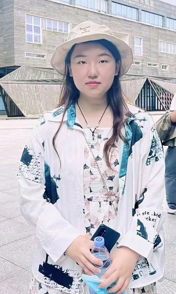 A young woman with long dark hair wearing a beige hat, a patterned shirt, and a white jacket, holding a smartphone, water bottle, and face mask, standing outdoors with a modern building in the background.