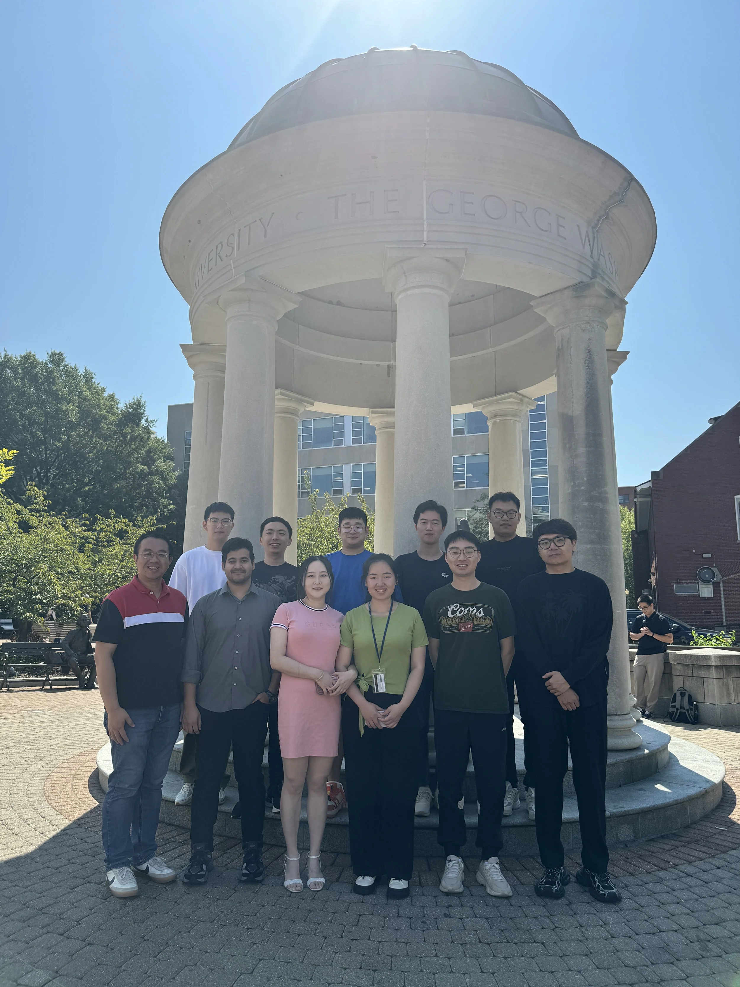 A group of eleven people standing in front of a historic monument with columns and a dome, with the text 'UNIVERSITY • THE GEORGE WASH' inscribed on it. The setting appears to be outdoors on a sunny day with trees and buildings in the background.