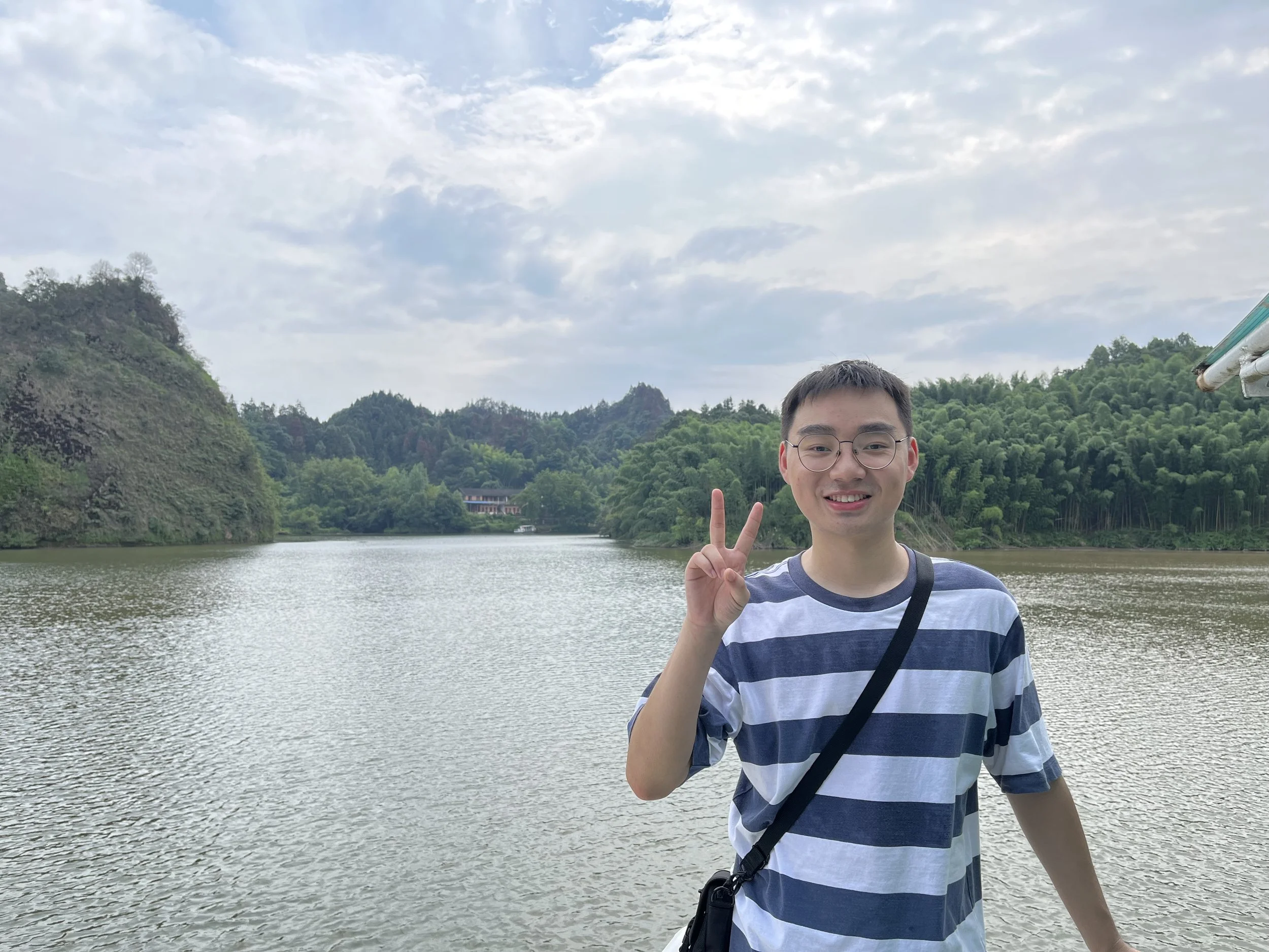 A young man with glasses and a striped t-shirt near a river, holding up peace signs with both hands, with green hills and a cloudy sky in the background.