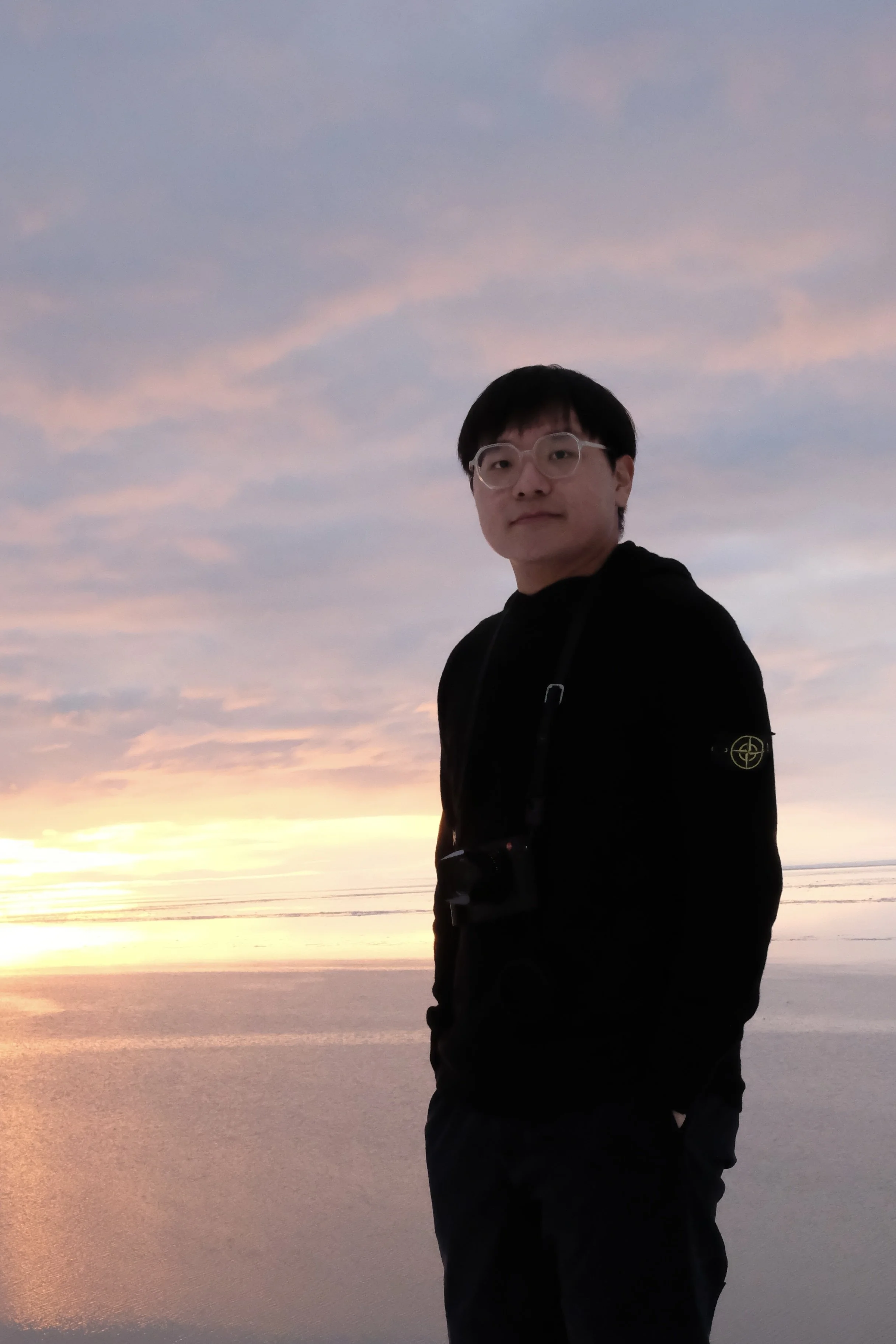 A person with black hair, glasses, and wearing a black jacket stands on a beach at sunset, with pink and purple clouds in the sky and calm water reflecting the colors.
