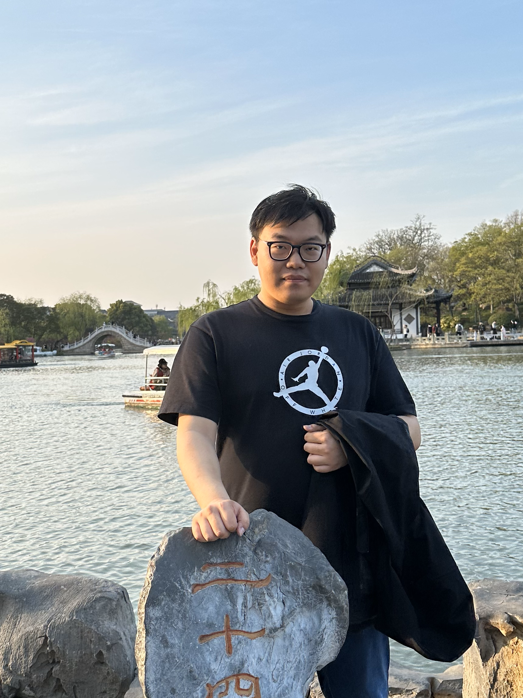 A man wearing glasses and a black T-shirt standing outdoors by a body of water with a traditional Chinese-style building and trees in the background. He is holding onto a large rock with carvings and symbols.