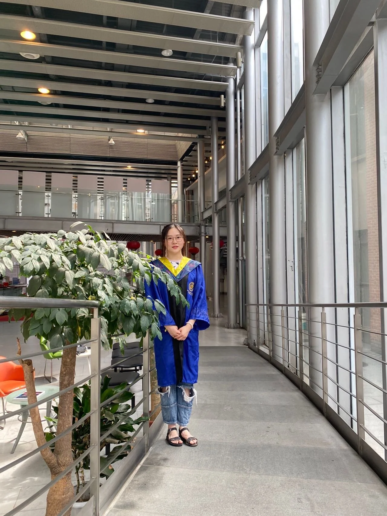 Young woman in a graduation cap and gown standing indoors near a glass wall and green plants.