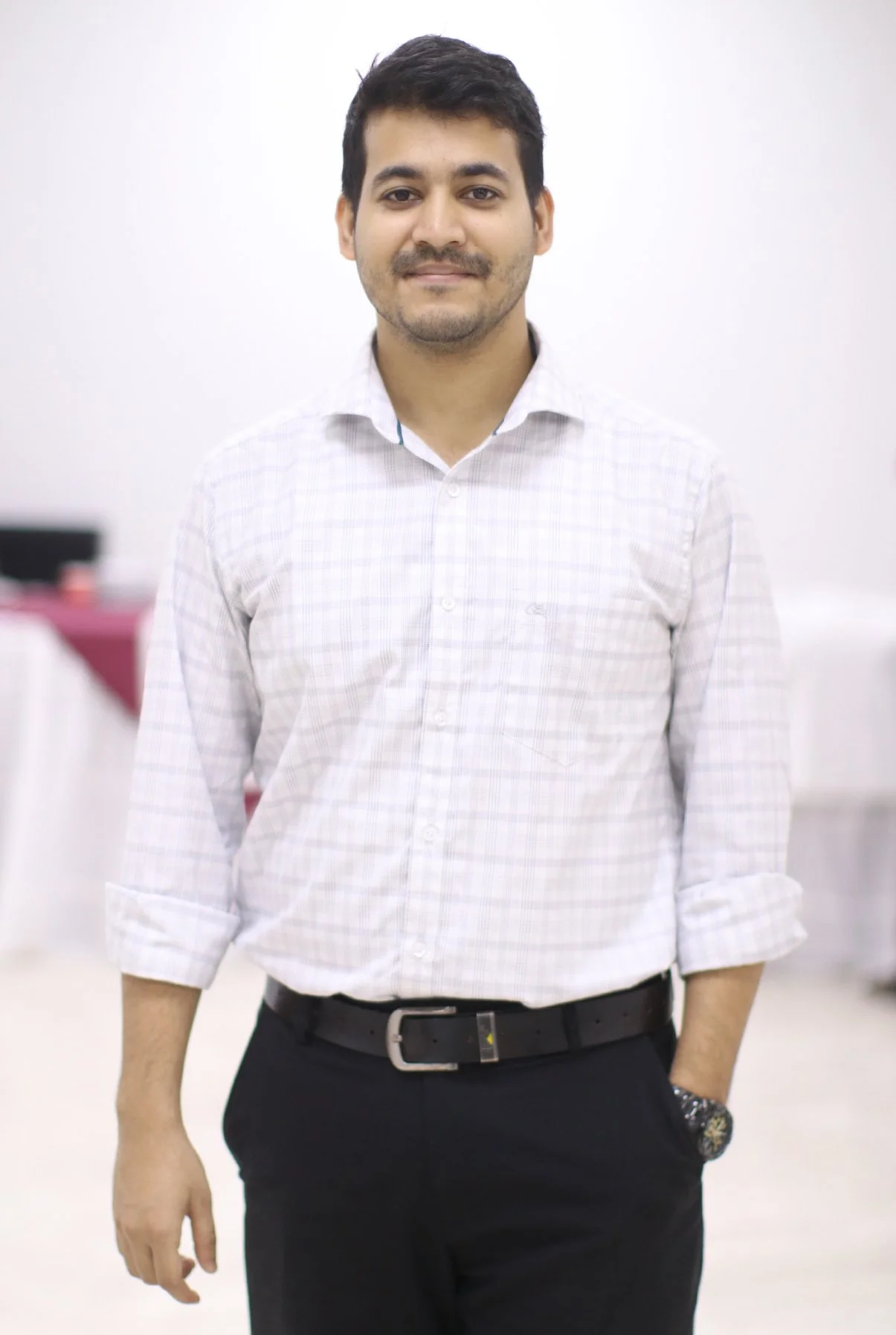 A young man with dark hair, light brown skin, and a slight beard, wearing a white checkered dress shirt and black pants, standing in a white room.