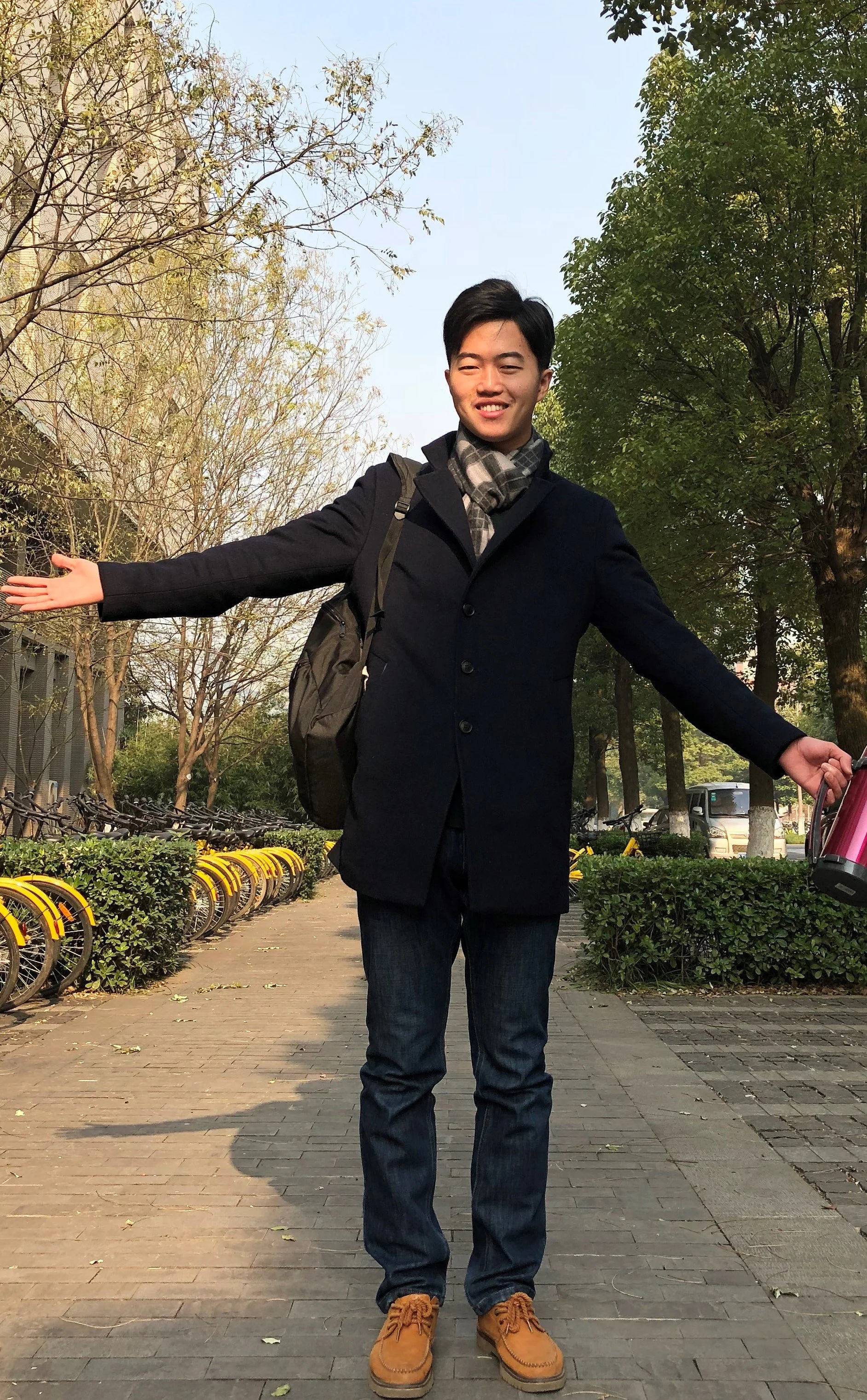 Young man with black hair smiling with arms open wide standing on a sidewalk in a park surrounded by trees, bicycles, and shrubs on a sunny day.