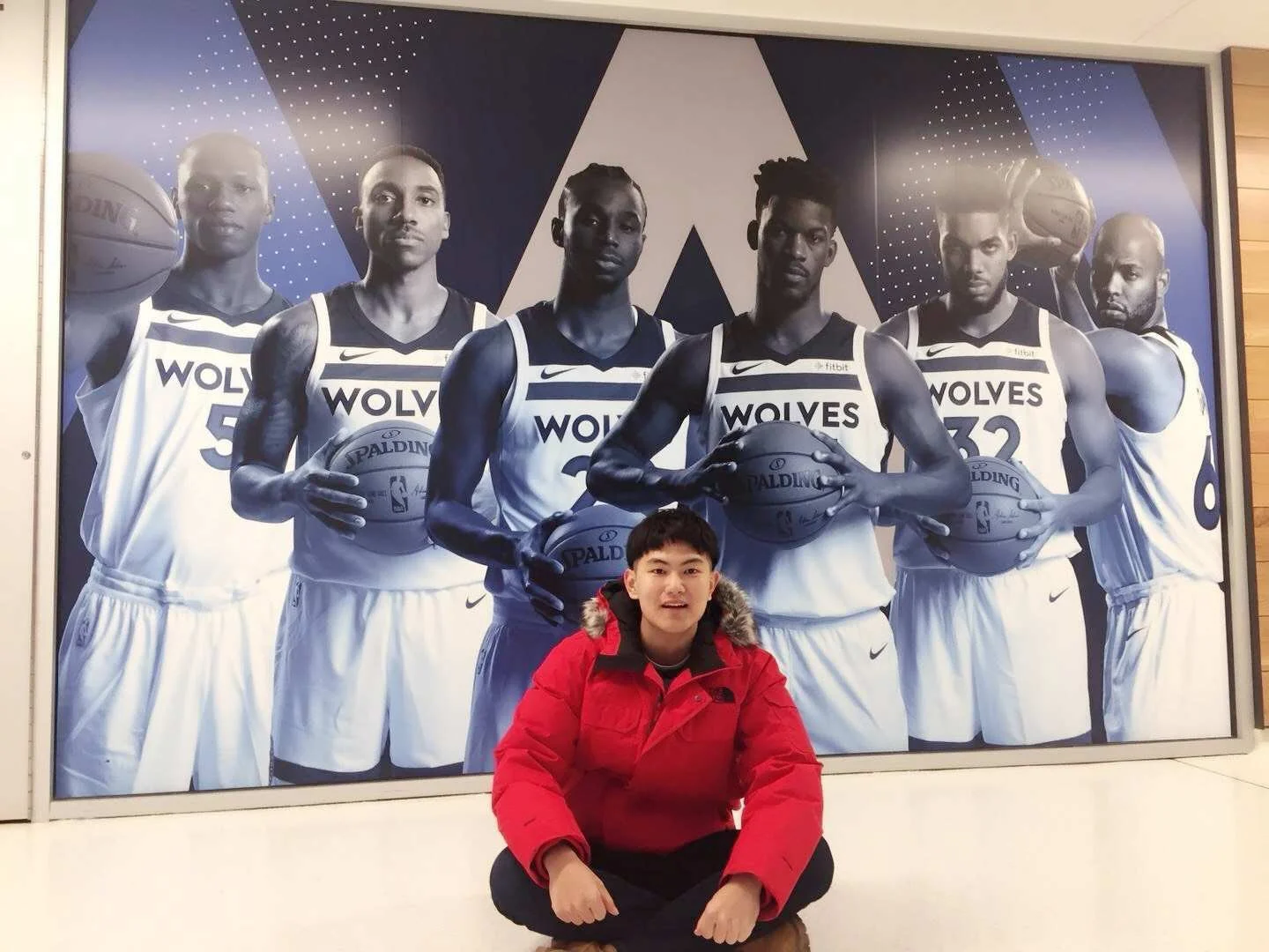 A person sitting cross-legged in front of a large black and white photograph of six basketball players wearing uniform with 'WOLVES' on them, holding basketballs. The person in the foreground is wearing a red jacket with fur-lined hood.