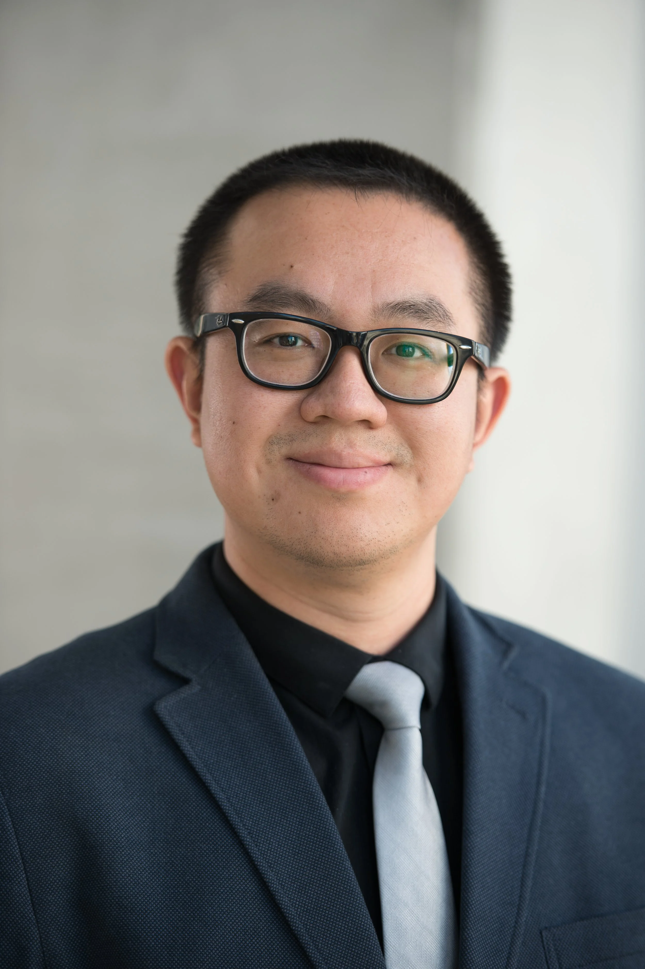 A young man with short black hair, wearing glasses, dressed in a dark suit, white shirt, and light gray tie, smiling in front of a plain light-colored background.
